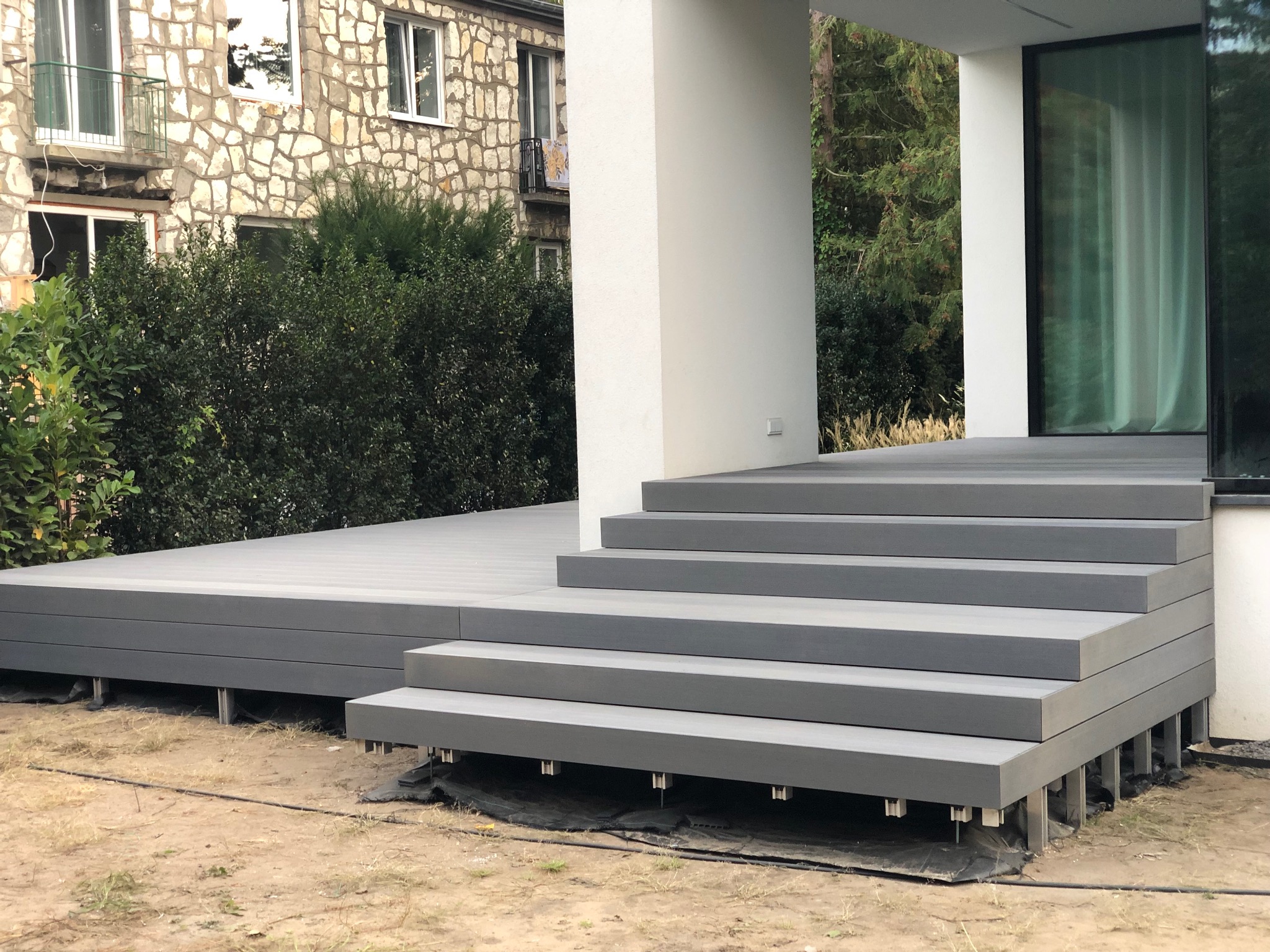 Nowoczesny, szary taras z kompozytu z czterema stopniami, wsparty na metalowych słupkach. W tle budynek z kamienia i zieleń.