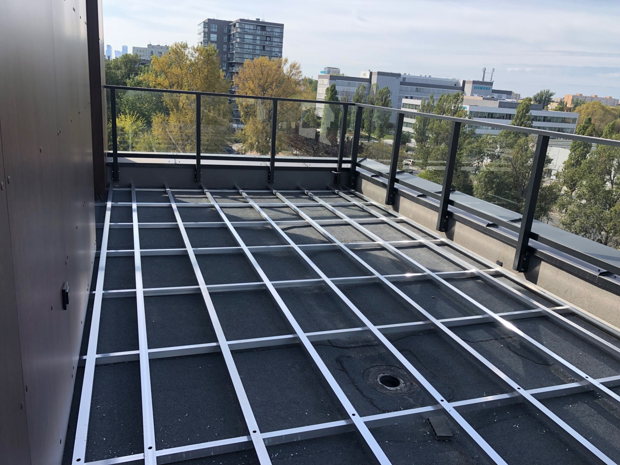 Montaż aluminiowej konstrukcji pod taras na dachu budynku z widokiem na miasto. Szara membrana dachowa i szklane balustrady. Etap budowy.