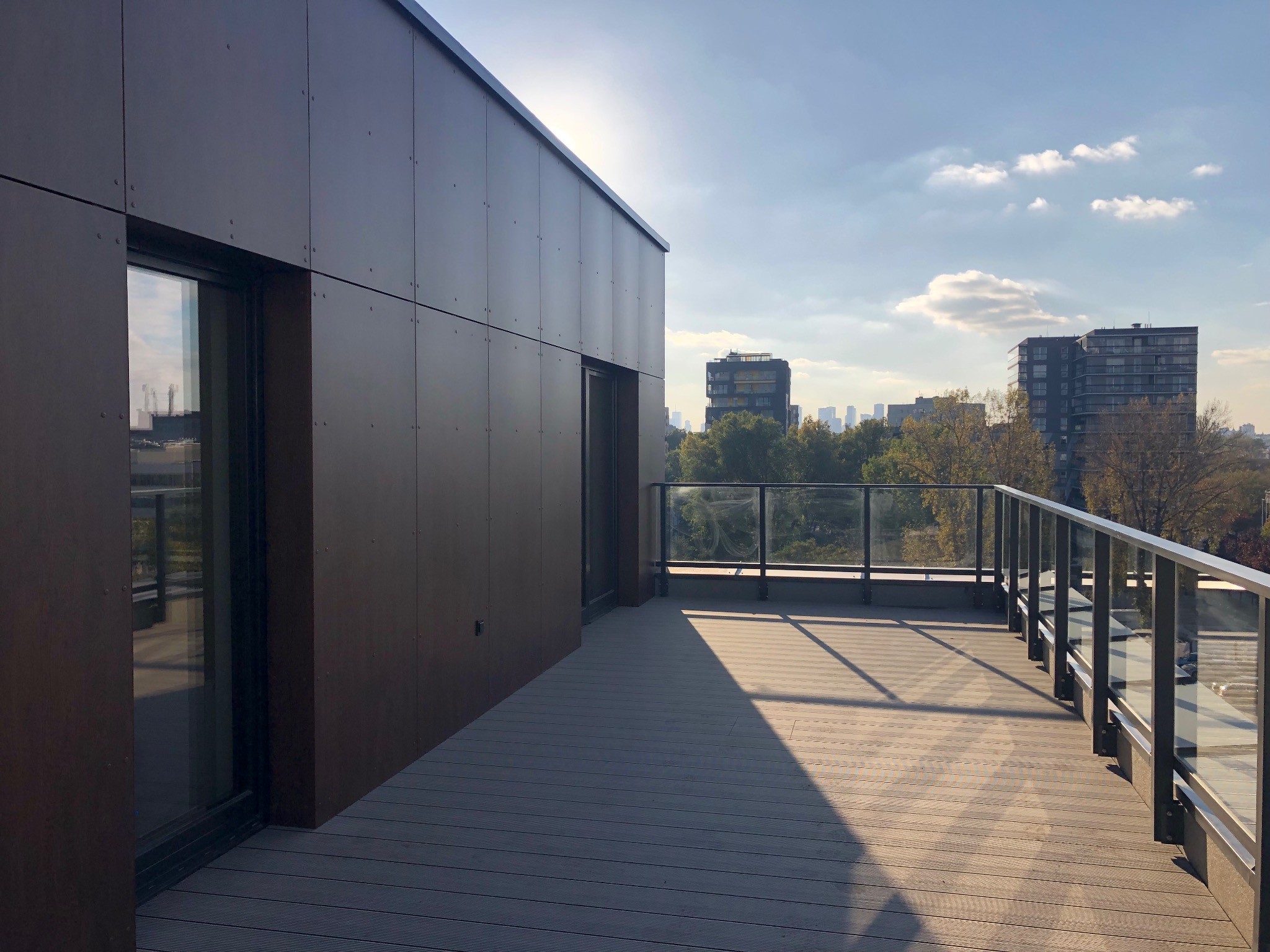 Nowoczesny taras na dachu budynku z szarych desek kompozytowych i szklaną balustradą, z widokiem na panoramę miasta w tle, oświetlony popołudniowym słońcem.