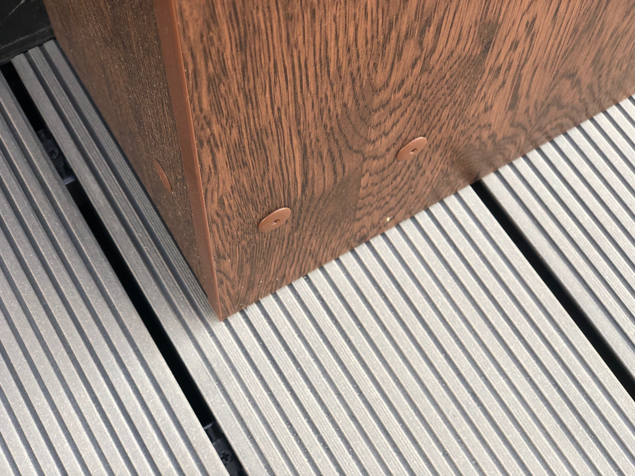 Detale wykończenia tarasu kompozytowego: brązowa donica z tworzywa sztucznego na szarej, ryflowanej desce tarasowej. Widoczne mocowania donicy.