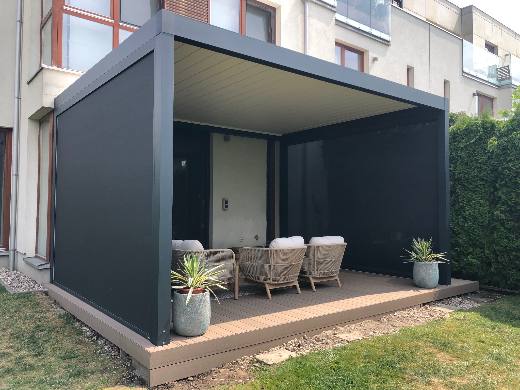 Nowoczesna, grafitowa pergola z roletami zaciemniającymi i tarasem kompozytowym. Na tarasie trzy fotele i donice z roślinami. Tło: budynek mieszkalny i trawnik.
