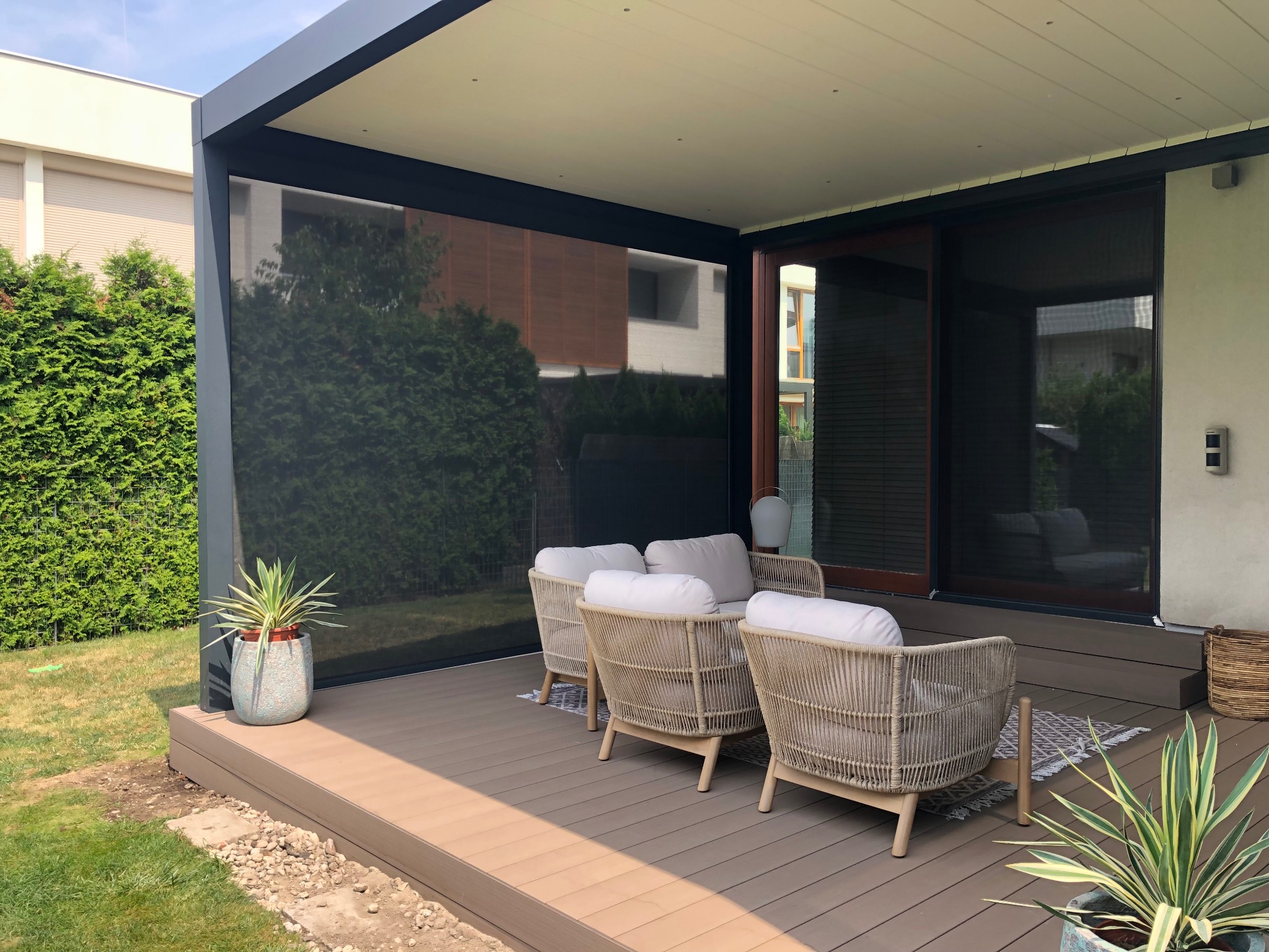 Zadaszony taras z kompozytowych desek w odcieniach brązu, wyposażony w rattanowe meble wypoczynkowe i rośliny doniczkowe. Nowoczesna konstrukcja z przesłoną.