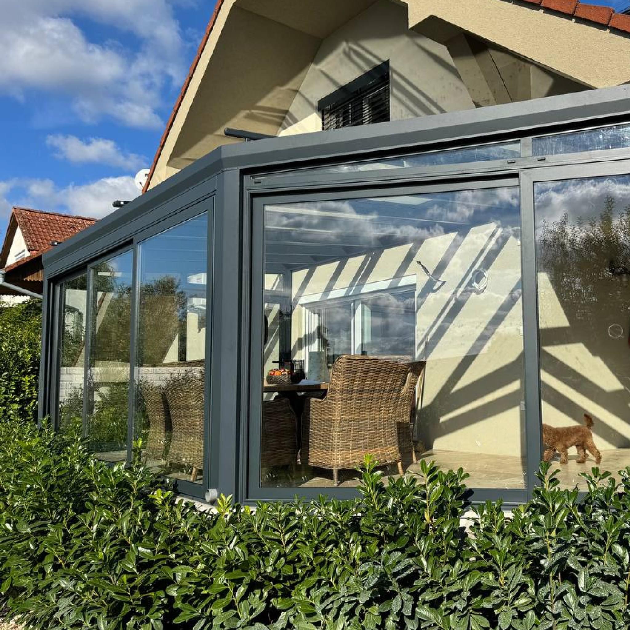 Szklana zabudowa tarasu z szarymi ramami, widok z zewnątrz na wiklinowe krzesła i stół z owocami. W tle dom z czerwoną dachówką i błękitne niebo.