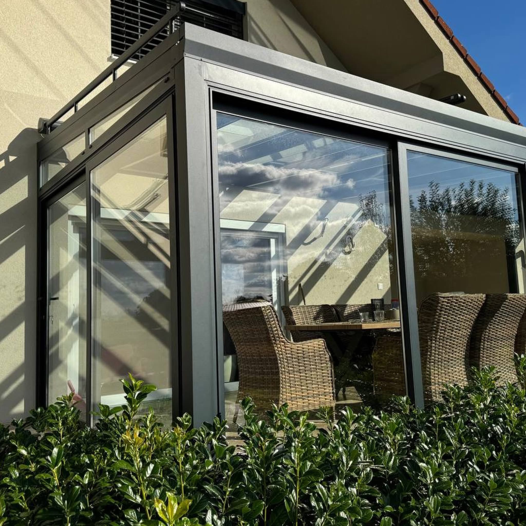 Szklana zabudowa tarasu z metalową ramą, widok z zewnątrz przez krzewy. Wewnątrz widoczne meble ogrodowe i odbicie nieba w szklanym dachu.