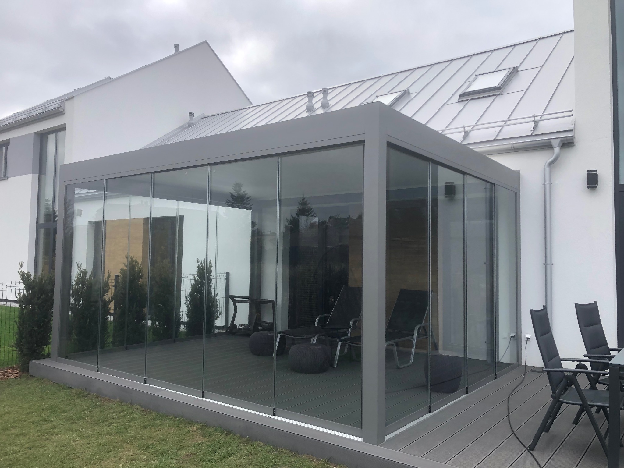 Szklana zabudowa tarasu z szarą ramą i meblami wypoczynkowymi wewnątrz, na tle nowoczesnego domu z szarą dachówką i zielonym trawnikiem.