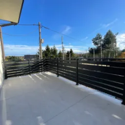 Czarna, pozioma balustrada na balkonie z szarymi płytkami, widok na las i błękitne niebo z chmurami.