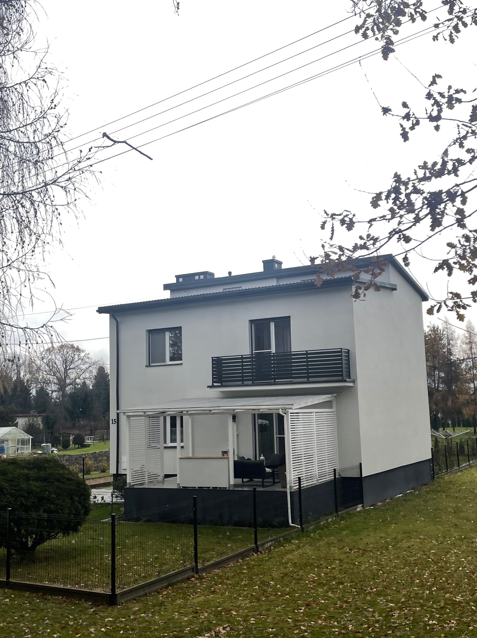 Nowoczesny dom z balkonem zabezpieczonym czarną barierką, białą pergolą i ogrodzeniem panelowym w kolorze czarnym.