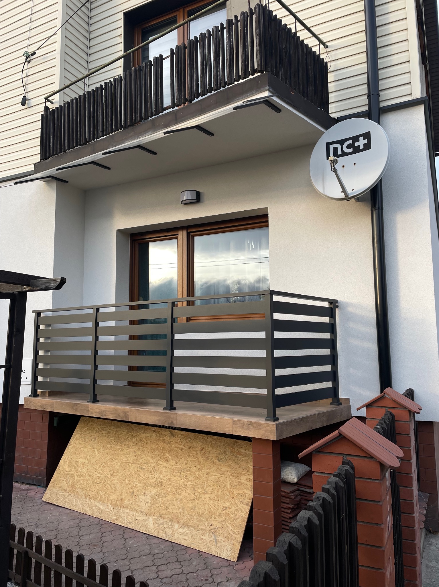 Balkon z nowoczesną, poziomą balustradą w kolorze grafitowym, widok z dołu, z widoczną anteną satelitarną na ścianie budynku i płytą OSB opartą o ścianę pod balkonem.