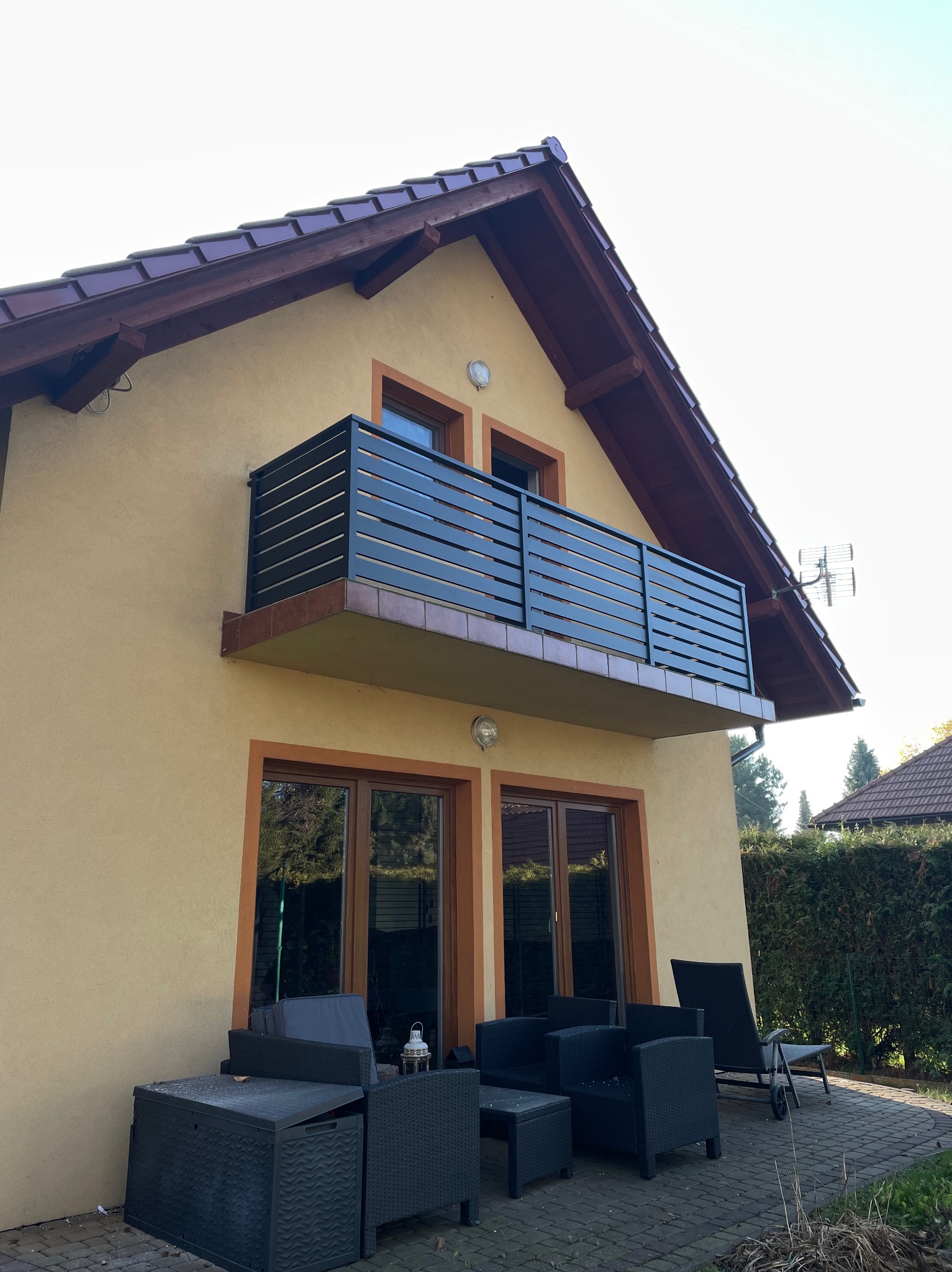 Czarna, pozioma balustrada balkonowa z ciemnego drewna, zamontowana na balkonie domu o żółtej elewacji, widok z dołu, meble ogrodowe na tarasie.