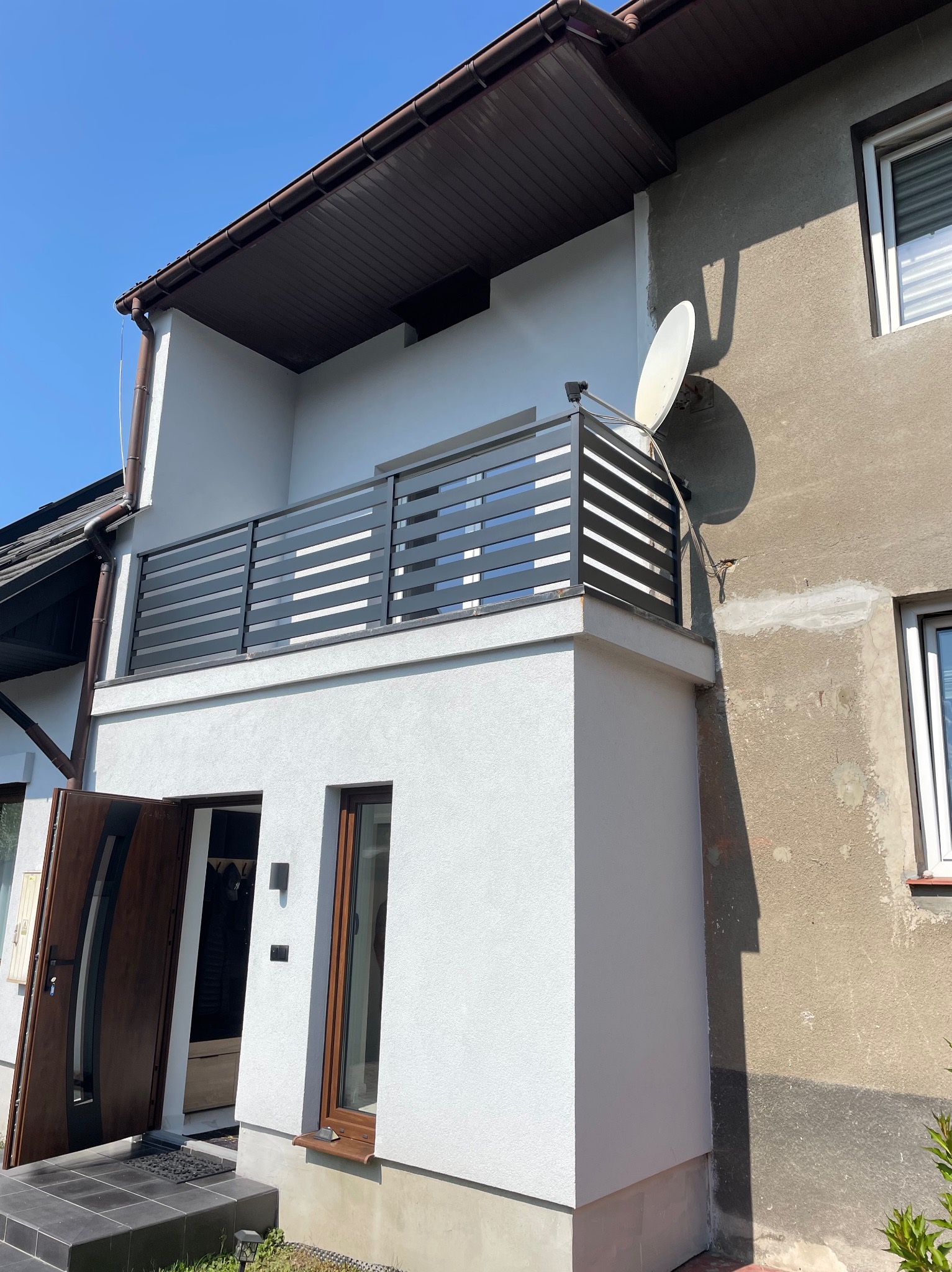 Nowoczesna balustrada balkonowa z poziomych profili w kolorze grafitowym, widok z dołu na tle błękitnego nieba i elewacji budynku z anteną satelitarną.