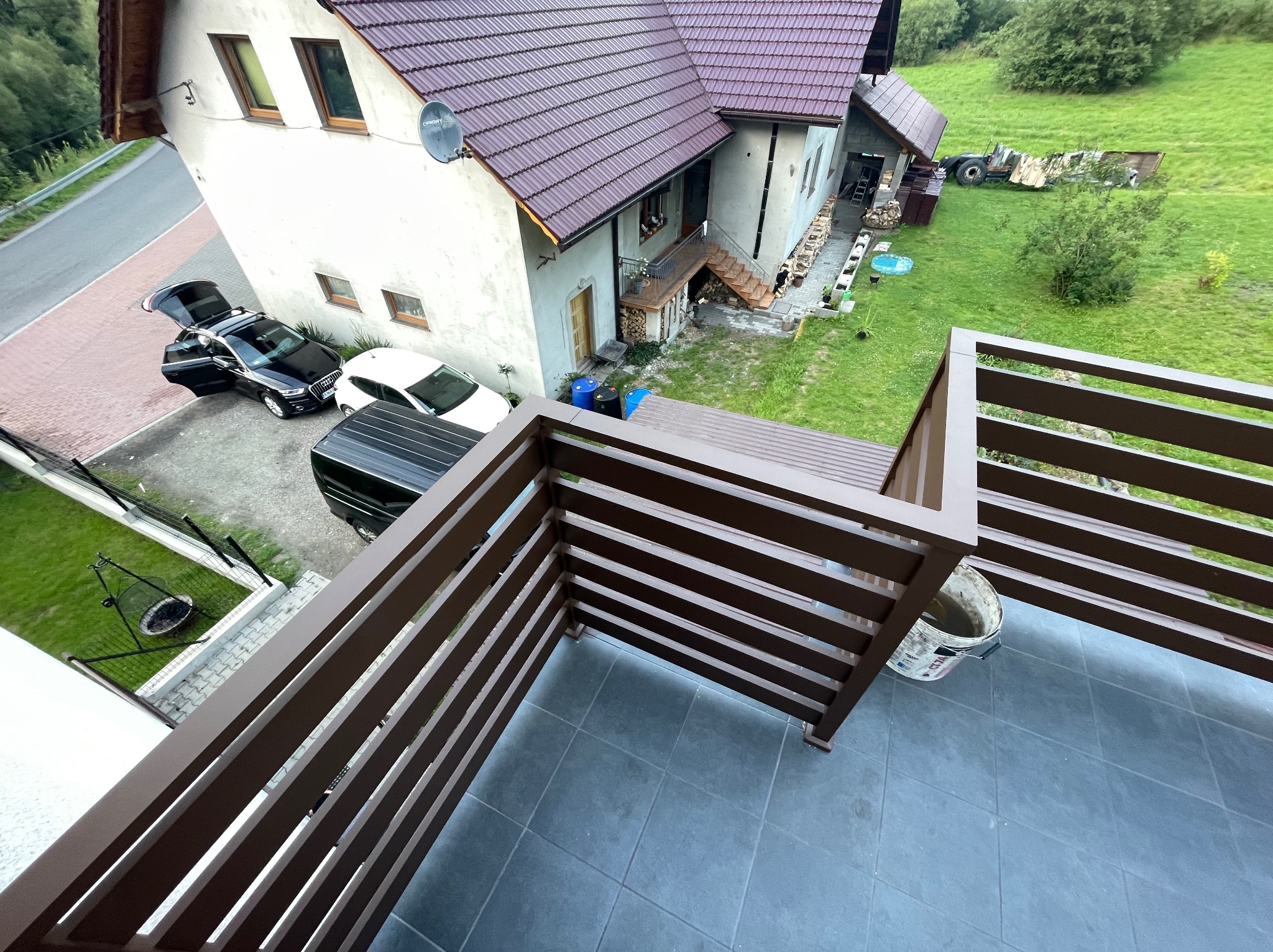 Balkon z brązową, poziomą balustradą z widokiem na dom z czerwonym dachem, samochody i zielone otoczenie.