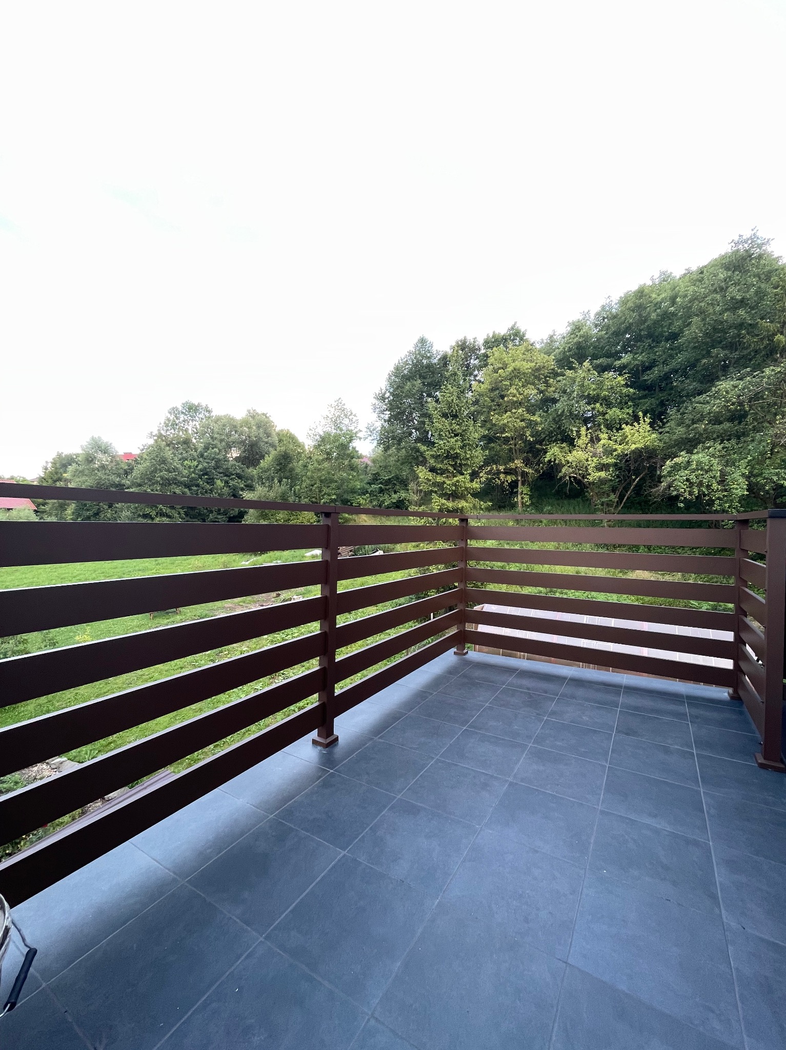 Balkon z brązową, poziomą balustradą z widokiem na zieleń drzew i łąkę, wykończony szarą, kwadratową terakotą.