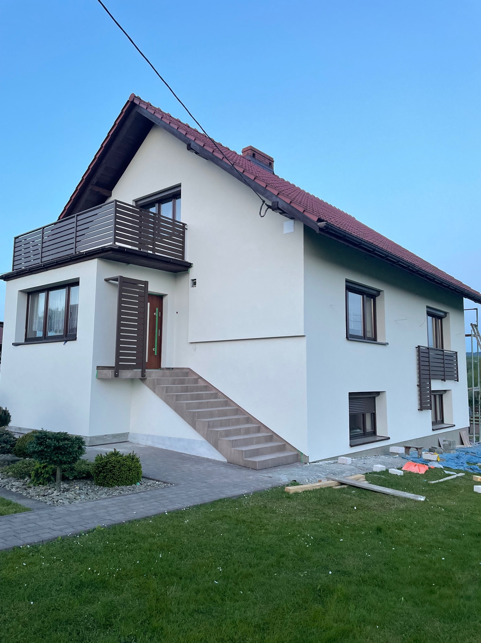 Nowoczesny dom jednorodzinny z brązowymi elementami wykończeniowymi, w tym balkon i pionowe elementy przy wejściu, widok z trawnika, częściowo widoczne materiały budowlane sugerują trwające prace.