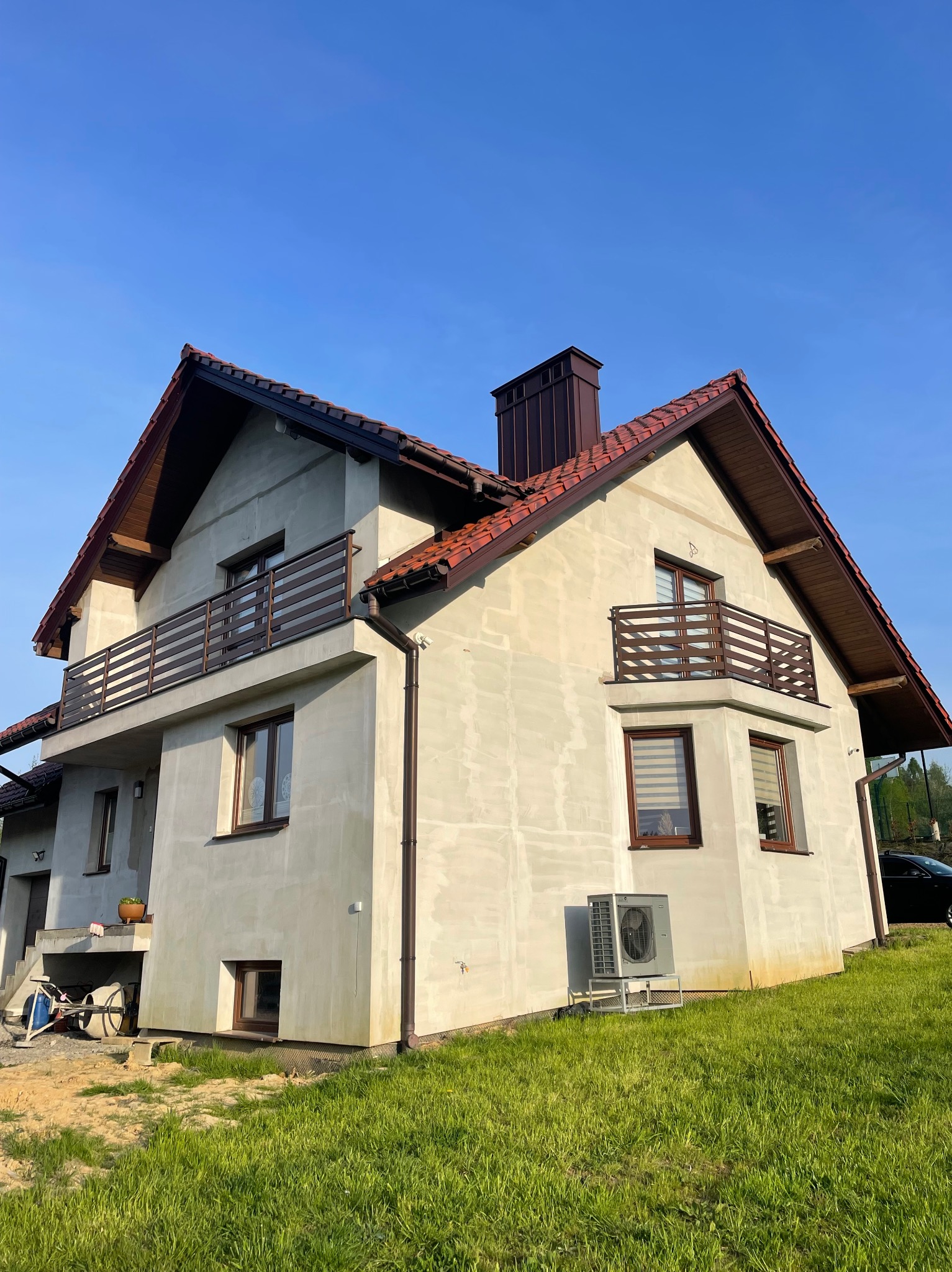 Dwupiętrowy dom w trakcie budowy z brązowymi, poziomymi barierkami balkonowymi i widocznym klimatyzatorem na trawniku.