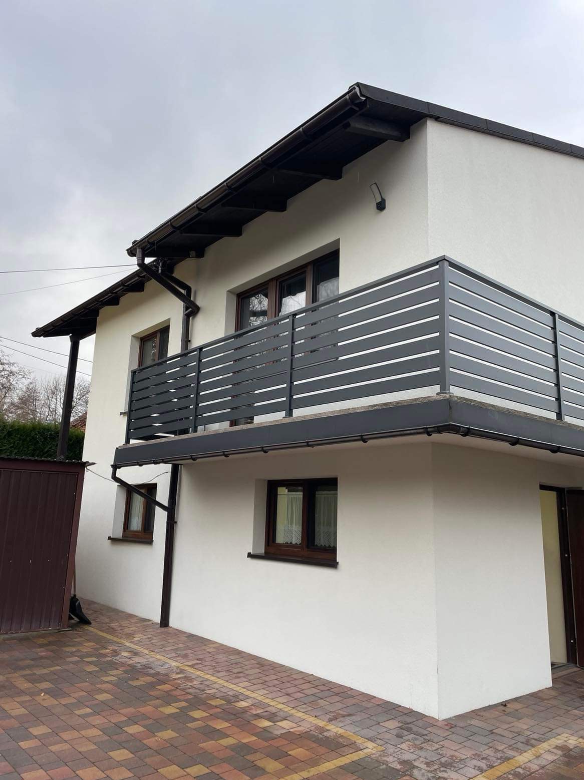 Nowoczesna balustrada balkonowa z poziomych profili w kolorze grafitowym, zamontowana na balkonie budynku z białą elewacją i brązowymi oknami.