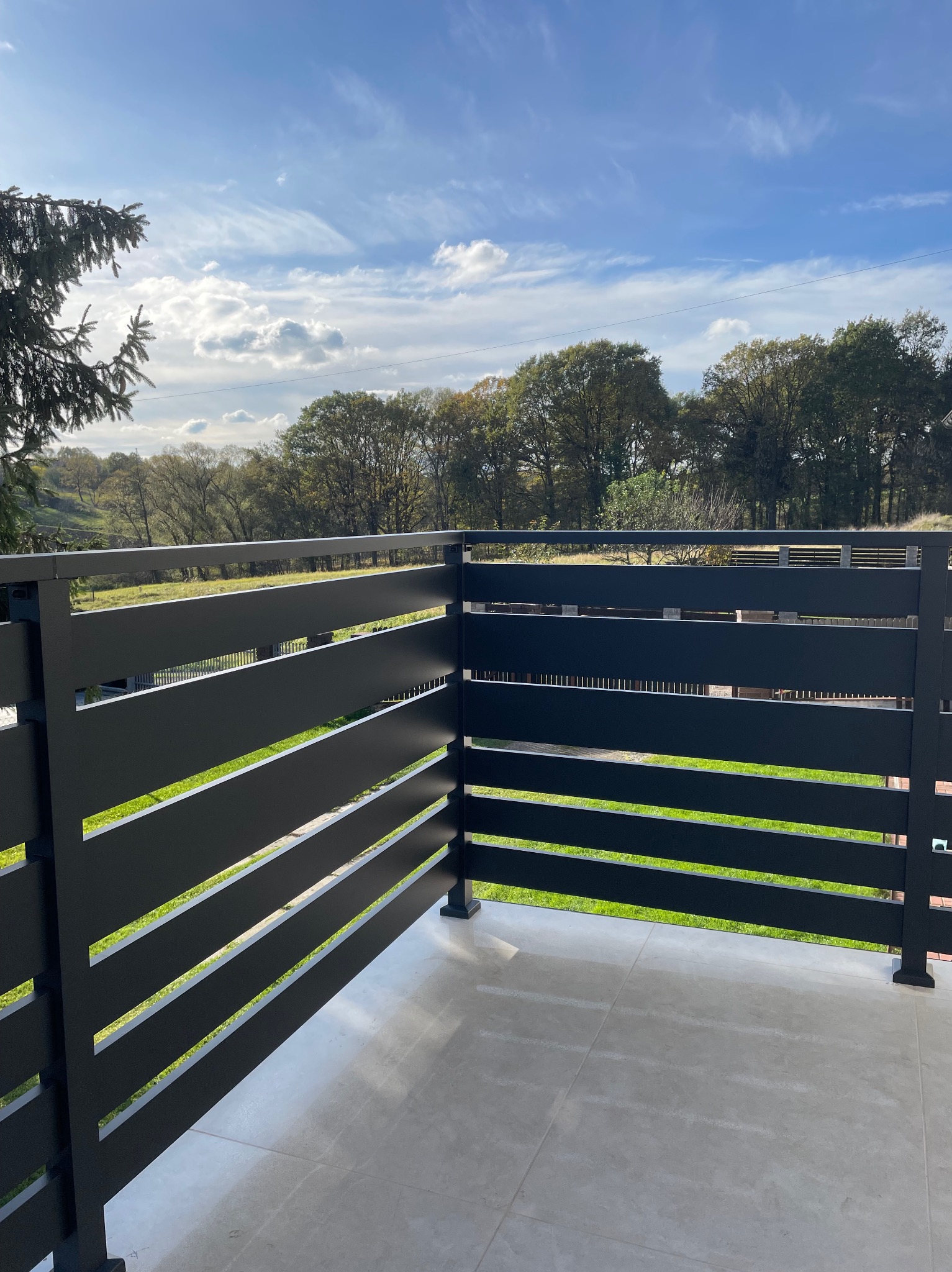 Nowoczesna, czarna balustrada balkonowa z poziomymi szczeblami, widok na zielony krajobraz i błękitne niebo z chmurami.