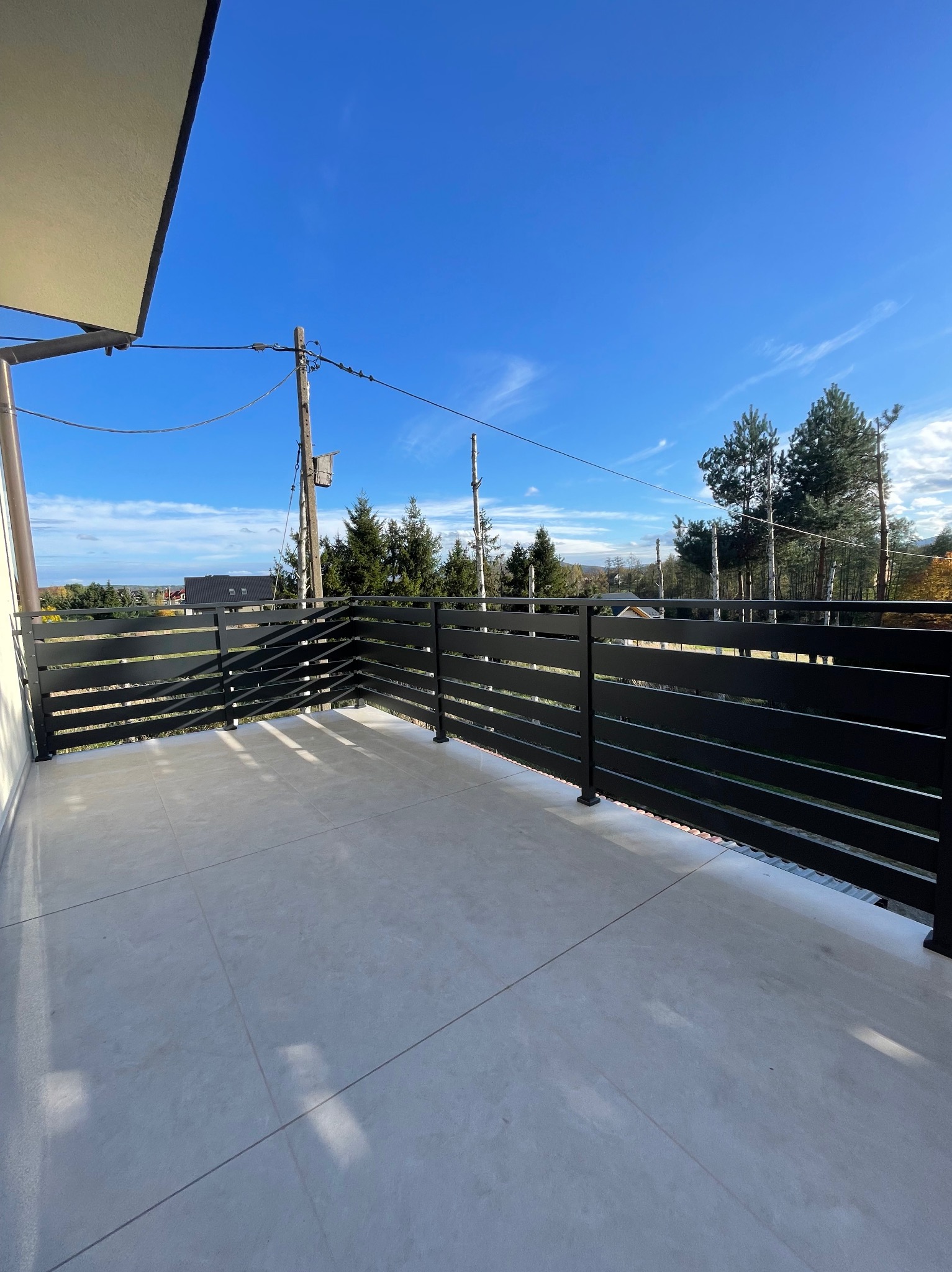 Czarna, pozioma balustrada na balkonie z szarymi płytkami, widok na las i błękitne niebo z chmurami.