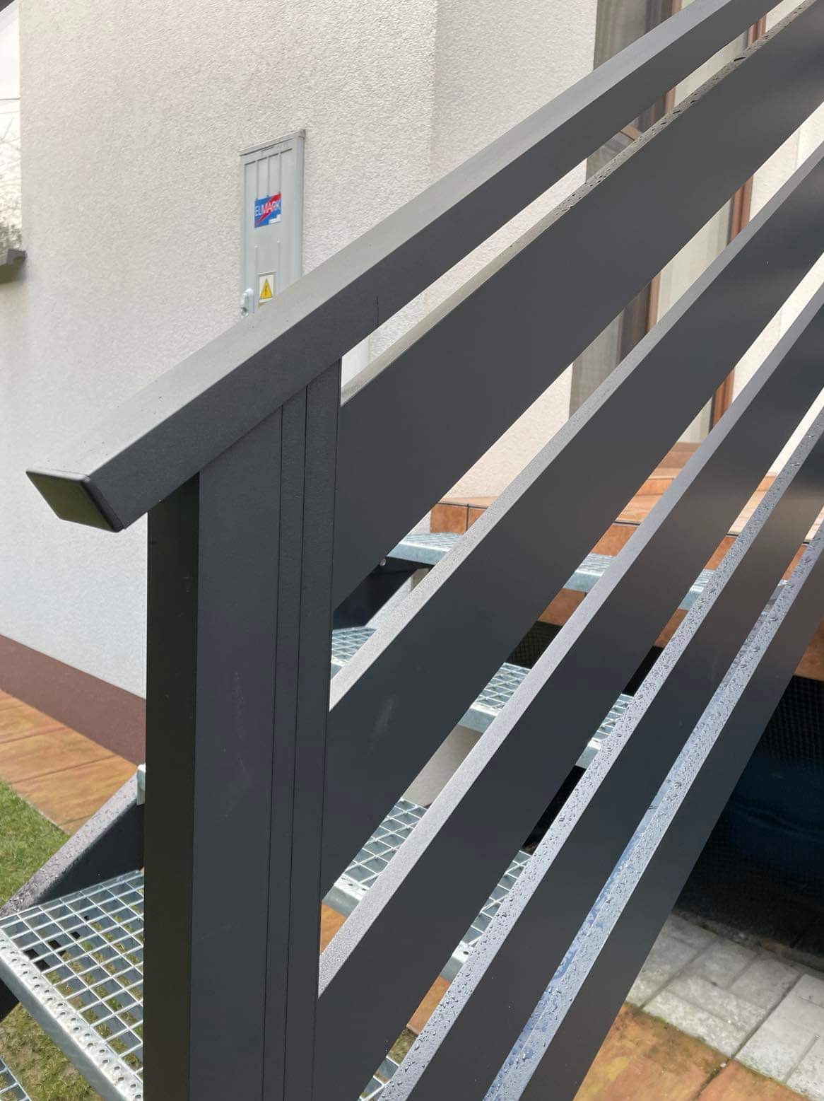 Metalowa poręcz schodowa w kolorze grafitowym, z pionowymi elementami, zamontowana przy zewnętrznych schodach z kraty pomostowej.