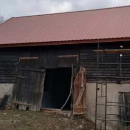 Stary drewniany budynek gospodarczy z nowym, metalowym dachem w kolorze ceglastym, widoczne rusztowanie i pracownik w odblaskowych spodniach. Dach pokryty blachą trapezową. Częściowo otwarte...