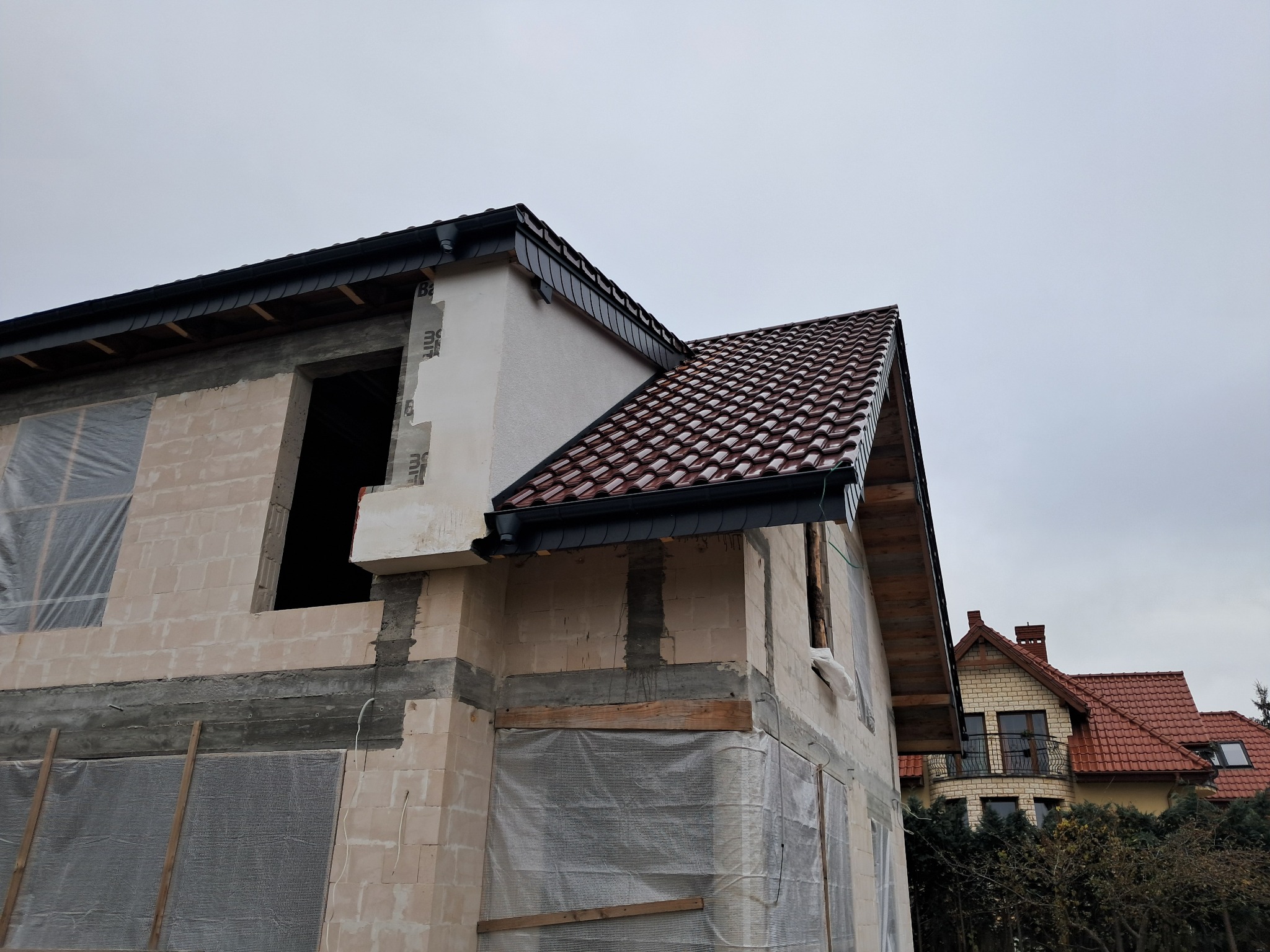 Surowy stan domu w trakcie budowy: widoczne mury z pustaków, częściowo zakryte folią, nowy dach z ciemnoczerwonej dachówki ceramicznej, czarne rynny, w tle dom z czerwonym dachem.