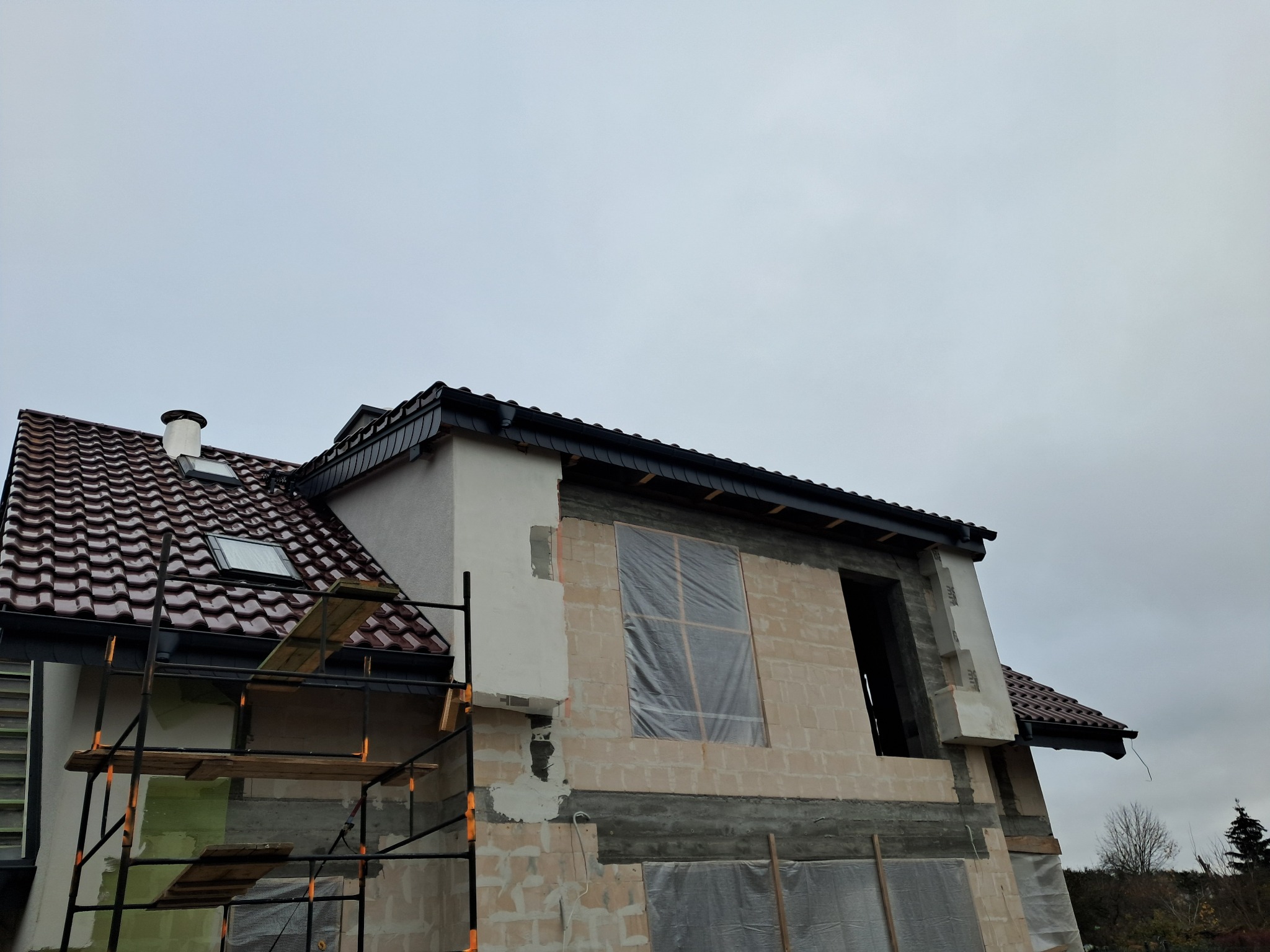 Dom w trakcie budowy z widocznymi rusztowaniami, częściowo pokryty dachem z dachówki ceramicznej, z oknem dachowym i fragmentem rynny, okno na wyższej kondygnacji zabezpieczone folią.