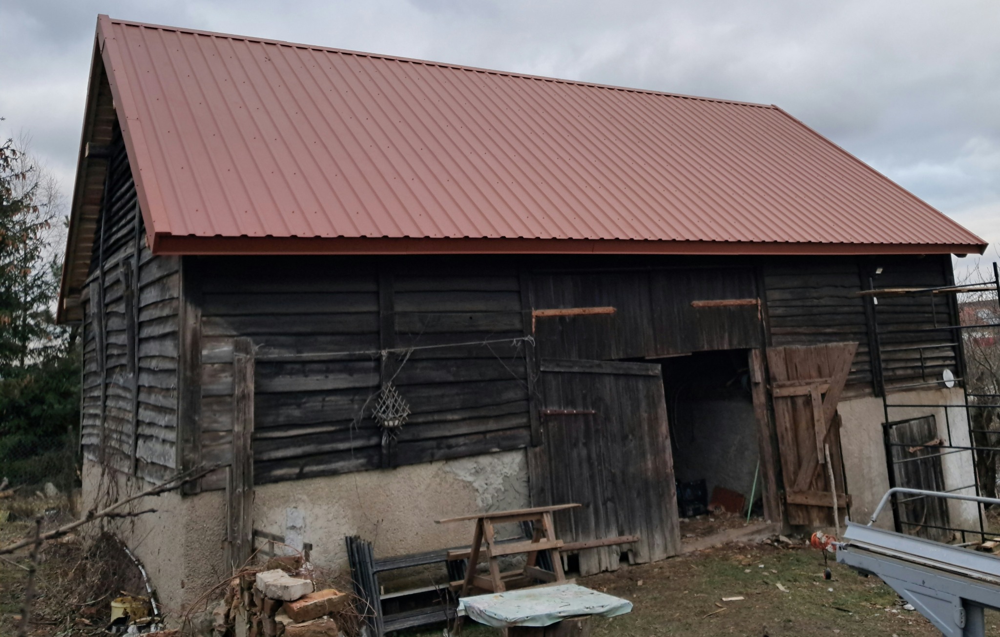 Stary, drewniany budynek gospodarczy z nowym, brązowym dachem z blachy trapezowej. Widoczne nierówności i patyna na drewnianej elewacji. Otwarta brama wjazdowa, a przed budynkiem porozrzucane...
