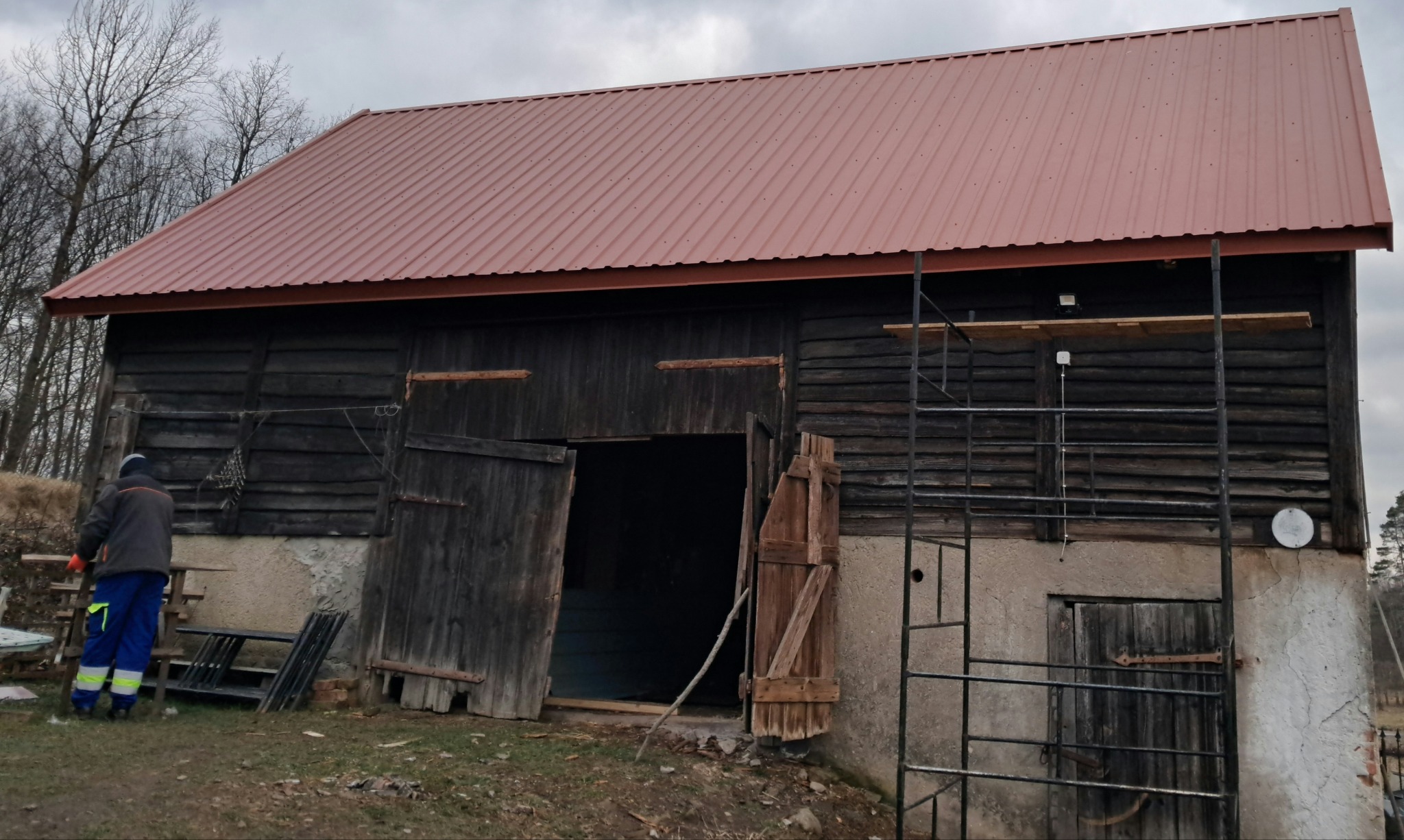 Stary drewniany budynek gospodarczy z nowym, metalowym dachem w kolorze ceglastym, widoczne rusztowanie i pracownik w odblaskowych spodniach. Dach pokryty blachą trapezową. Częściowo otwarte...