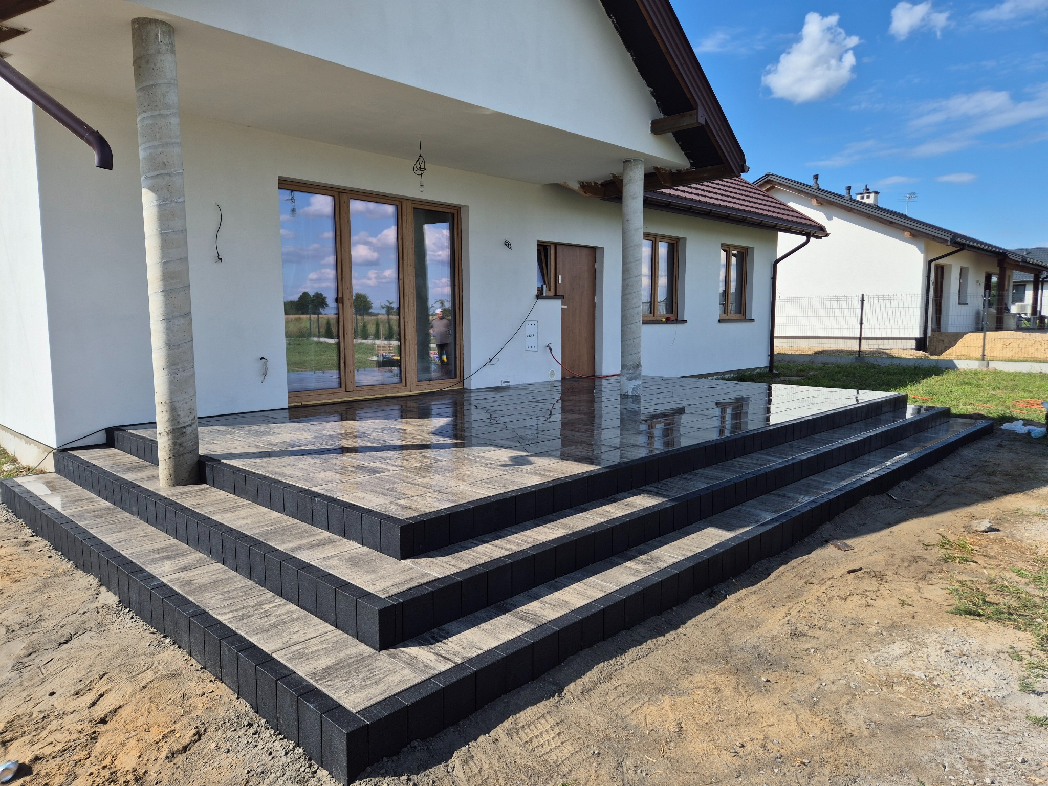 Ułożone schody z kostki brukowej prowadzące do domu jednorodzinnego. Ciemne obrzeża, jasne wypełnienie. Widoczne kolumny podtrzymujące zadaszenie.