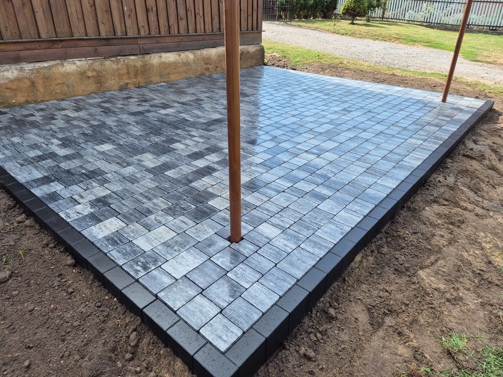 Świeżo ułożona, szara kostka brukowa w kwadratowy wzór, obramowana ciemną kostką. Widoczny fragment drewnianego budynku w tle i brązowy słupek przechodzący przez bruk.