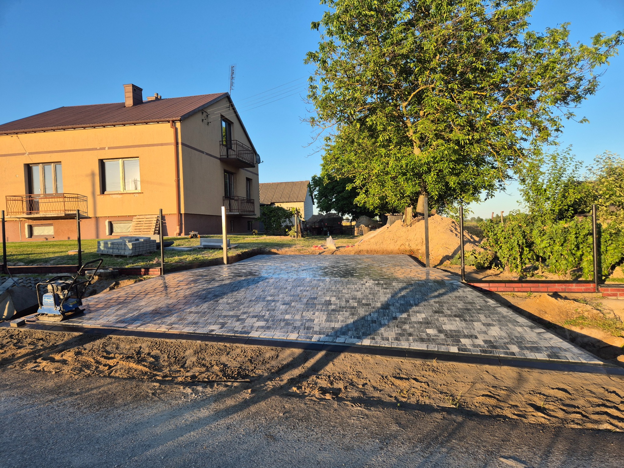 Częściowo ułożona, szara kostka brukowa na podjeździe przed domem jednorodzinnym, z widoczną zagęszczarką i przygotowanym terenem pod dalsze prace.