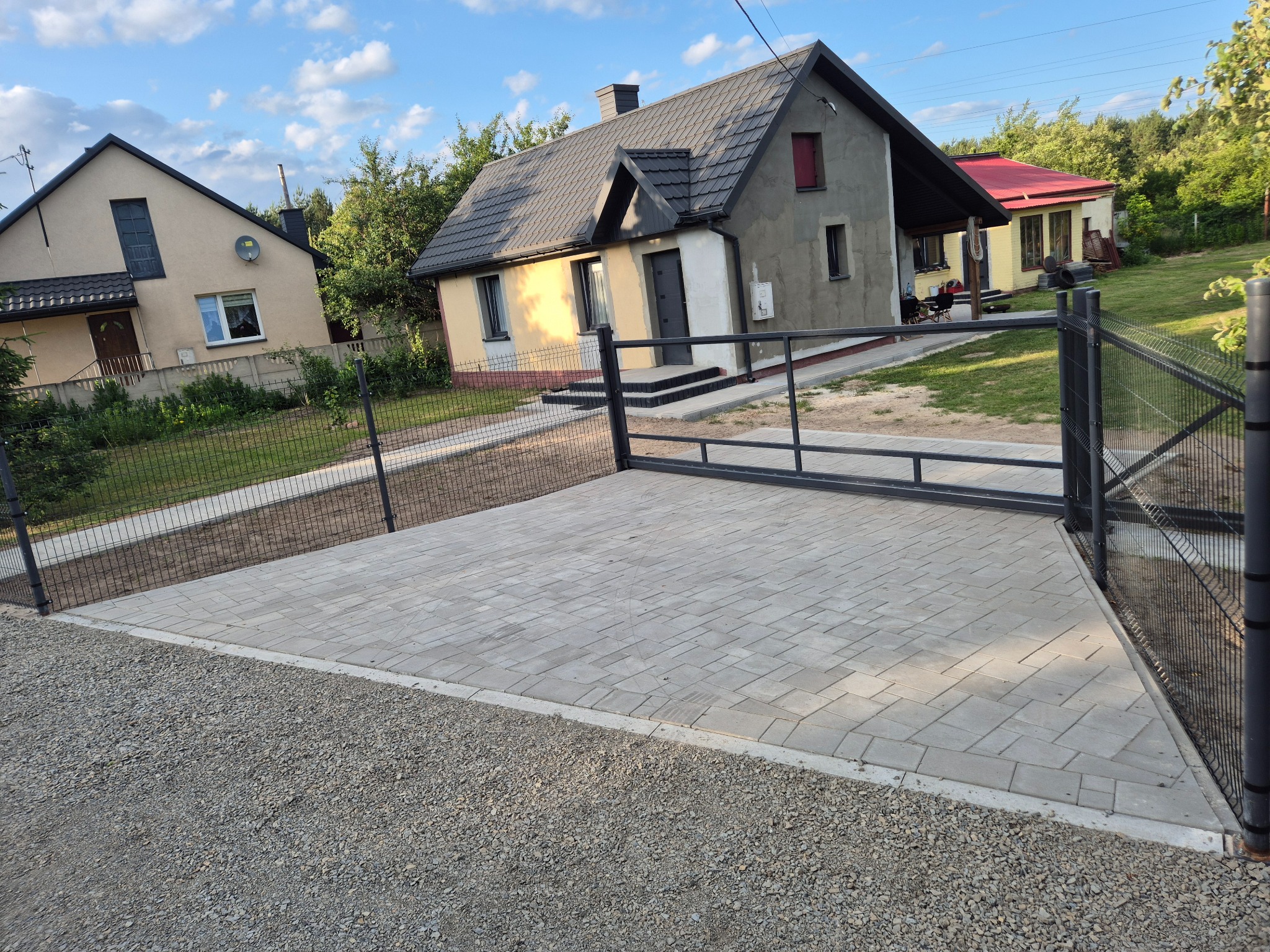 Nowo ułożona, szara kostka brukowa na podjeździe przed domem, z widoczną bramą w tle i drobnym żwirem po bokach. Słoneczny dzień.