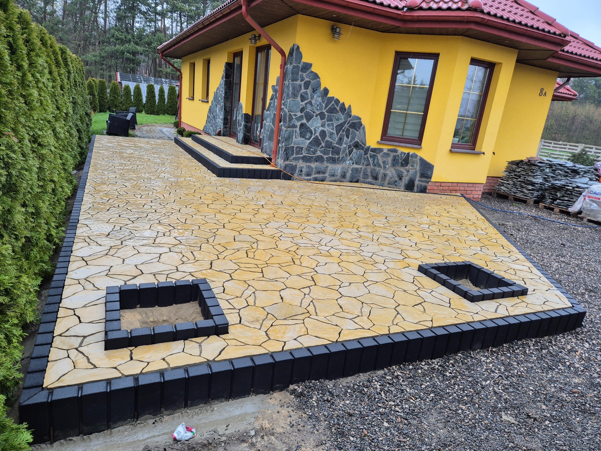 Żółta kostka brukowa o nieregularnym kształcie, obramowana czarnym obrzeżem, tworząca taras przed żółtym domem z elementami kamiennymi na elewacji.