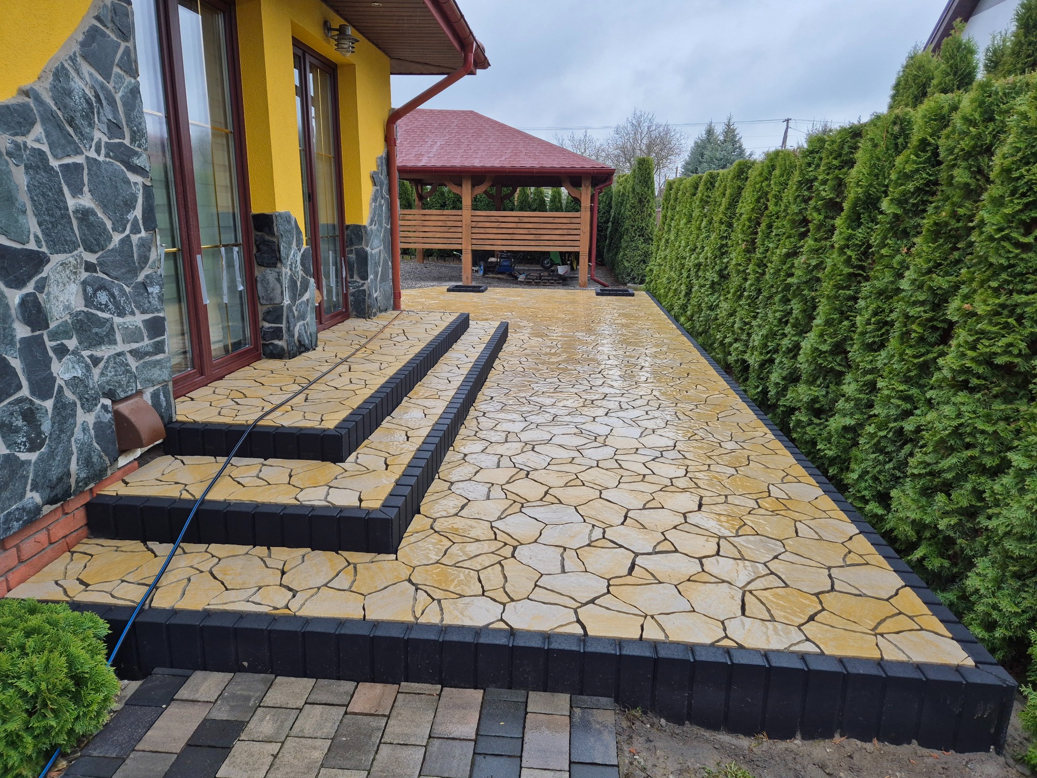 Żółta ścieżka z kamienia naturalnego z czarną obwódką prowadzi od domu do altany, obok żywopłot z tui. Widok na posesję w pochmurny dzień.