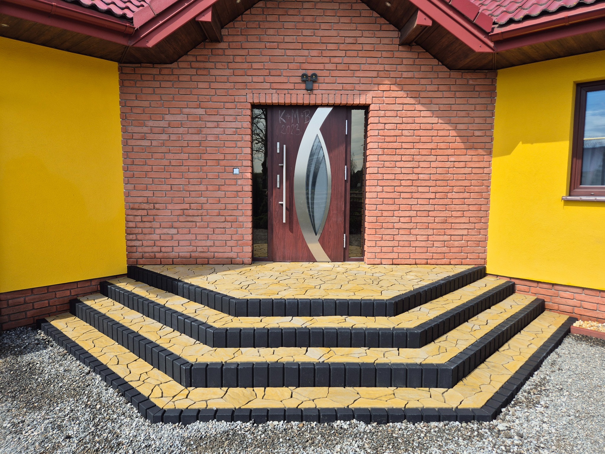 Schody wejściowe wyłożone żółto-czarną kostką brukową prowadzące do drzwi domu z cegły i żółtymi ścianami. Widok na ułożoną kostkę i wejście do budynku.