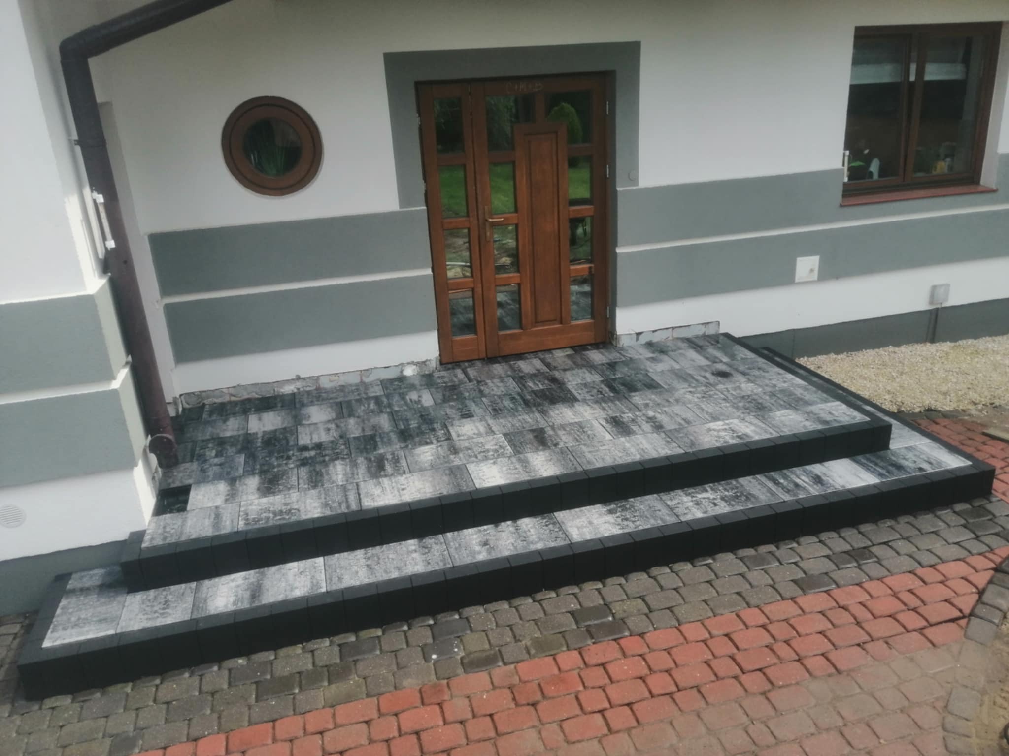 Schody wejściowe wyłożone szarą kostką brukową z czarnym obramowaniem, prowadzące do drewnianych drzwi wejściowych domu z oknem w tle i okrągłym oknem na górze.