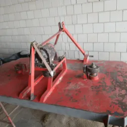 Czerwona kosiarka bijakowa z widocznymi śladami użytkowania i odpryskami farby, stojąca na betonowej podłodze przed ścianą z białych płytek.