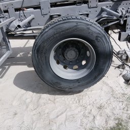 Karolina Świercz - Szare podwozie ciężarówki z nową oponą Michelin X MultiWay 3D na piaszczystym podłożu. Widoczne ślady stóp i trawa. Elementy podwozia po piaskowaniu.