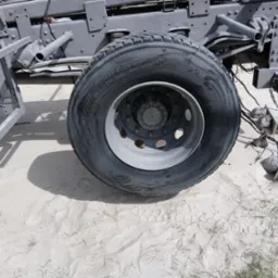 Szare podwozie ciężarówki z nową oponą Michelin X MultiWay 3D na piaszczystym podłożu. Widoczne ślady stóp i trawa. Elementy podwozia po piaskowaniu.