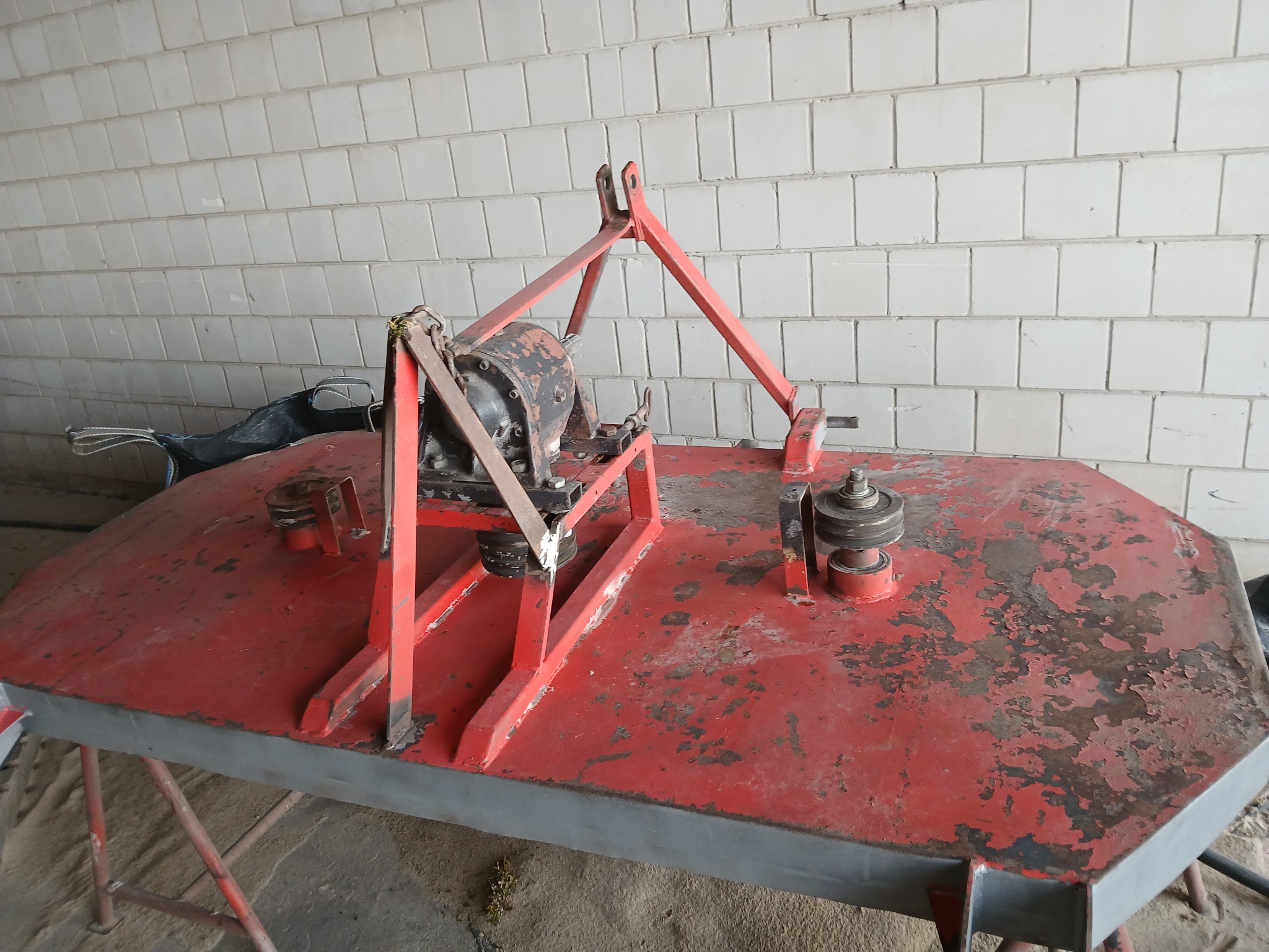 Czerwona kosiarka bijakowa z widocznymi śladami użytkowania i odpryskami farby, stojąca na betonowej podłodze przed ścianą z białych płytek.