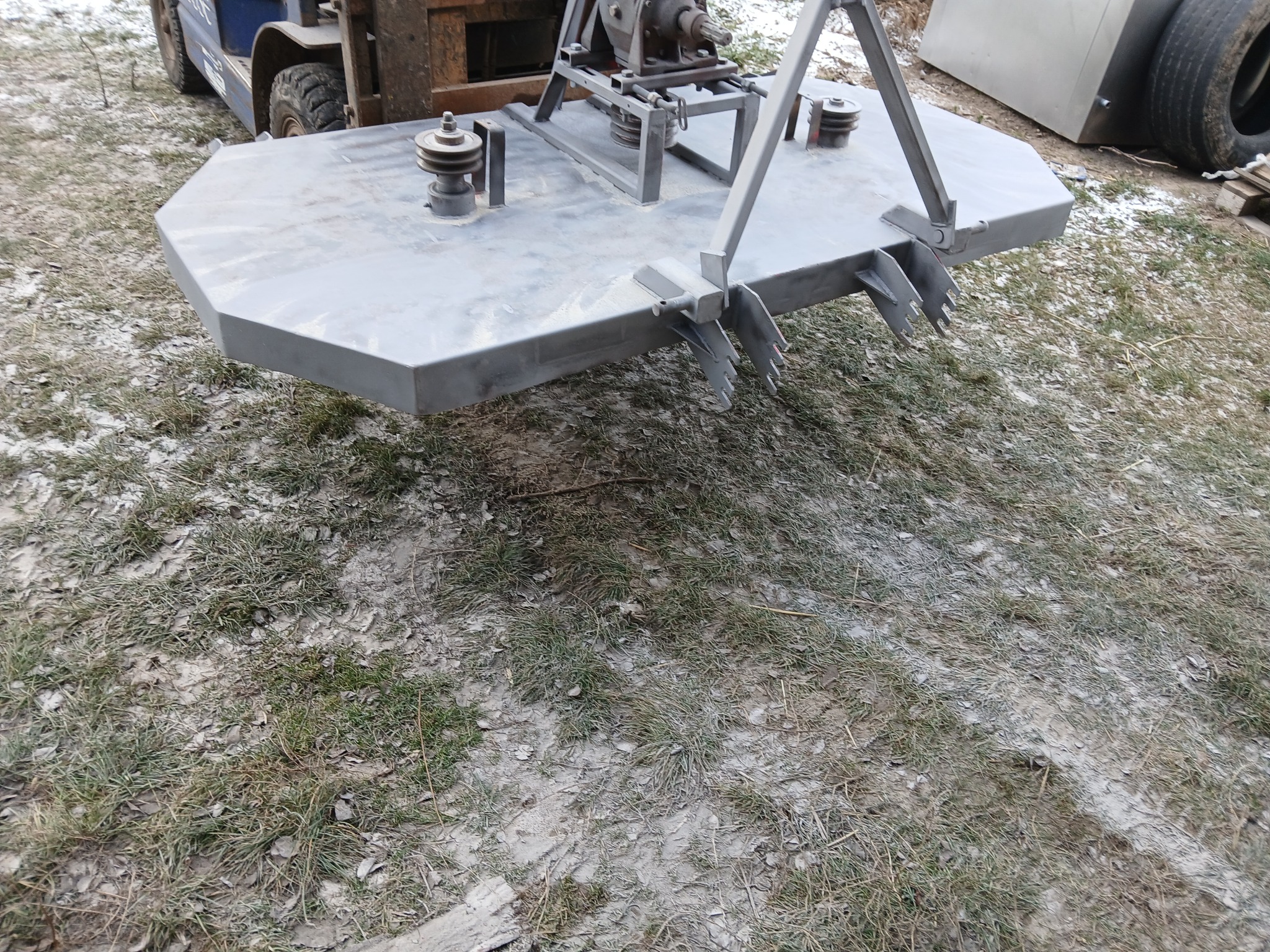 Szara kosiarka bijakowa zawieszona na wózku widłowym, gotowa do pracy na trawniku z resztkami śniegu.