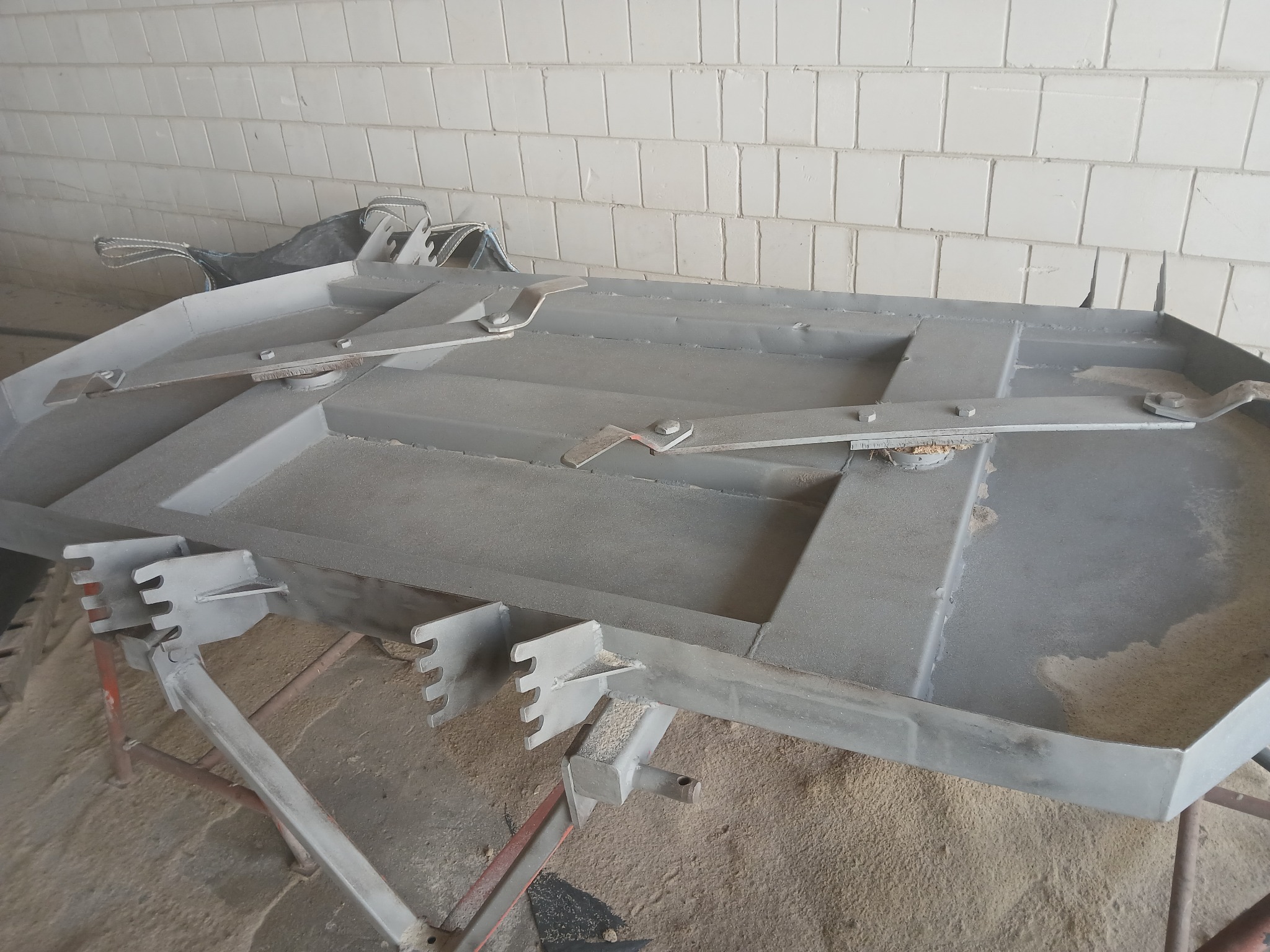 Szara kosiarka bijakowa po piaskowaniu, leżąca na stojaku w pomieszczeniu z białymi kafelkami w tle.