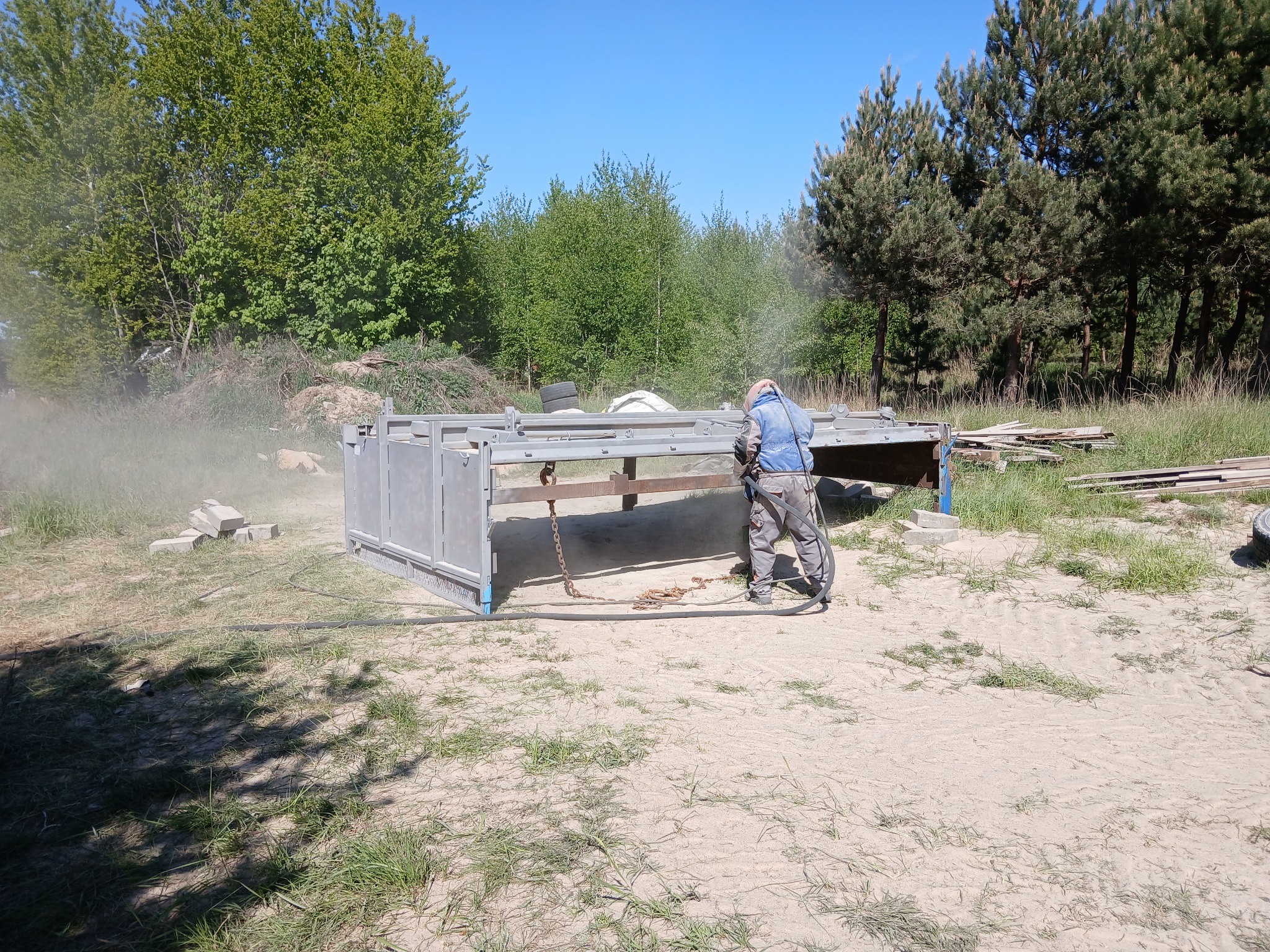 Pracownik w kombinezonie piaskuje metalową konstrukcję na zewnątrz, w tle drzewa i błękitne niebo. Widoczne efekty piaskowania.