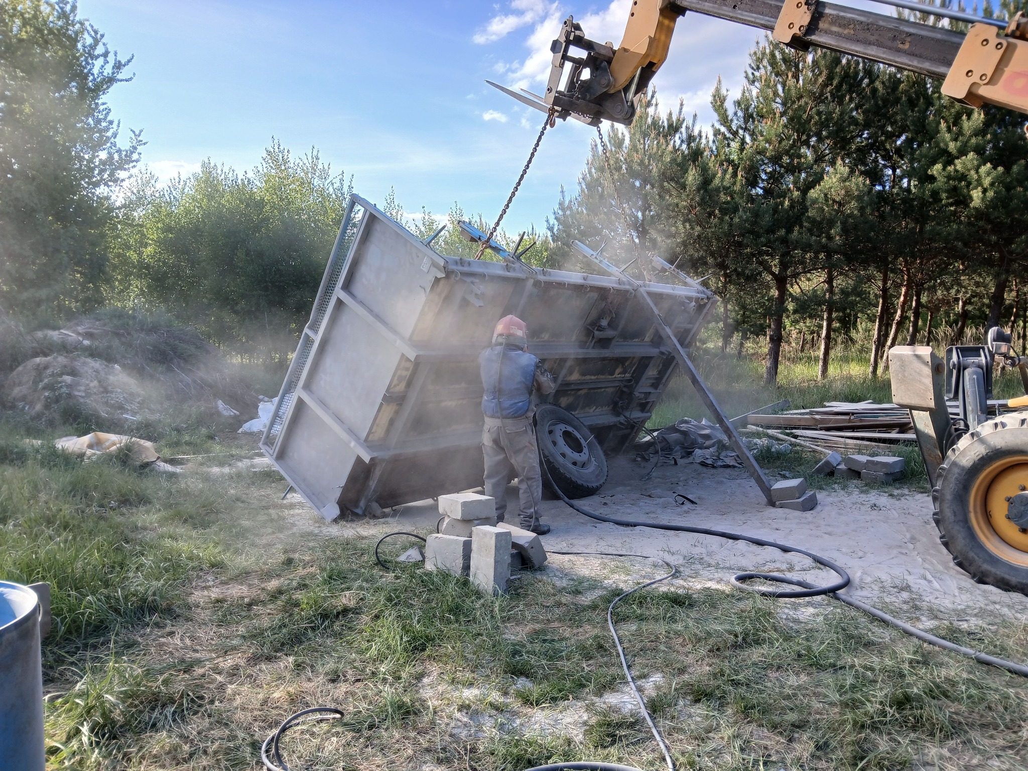 Operator w kasku piaskuje metalowy kontener podwieszony na łańcuchach przez maszynę budowlaną. Widoczne drzewa i trawa.