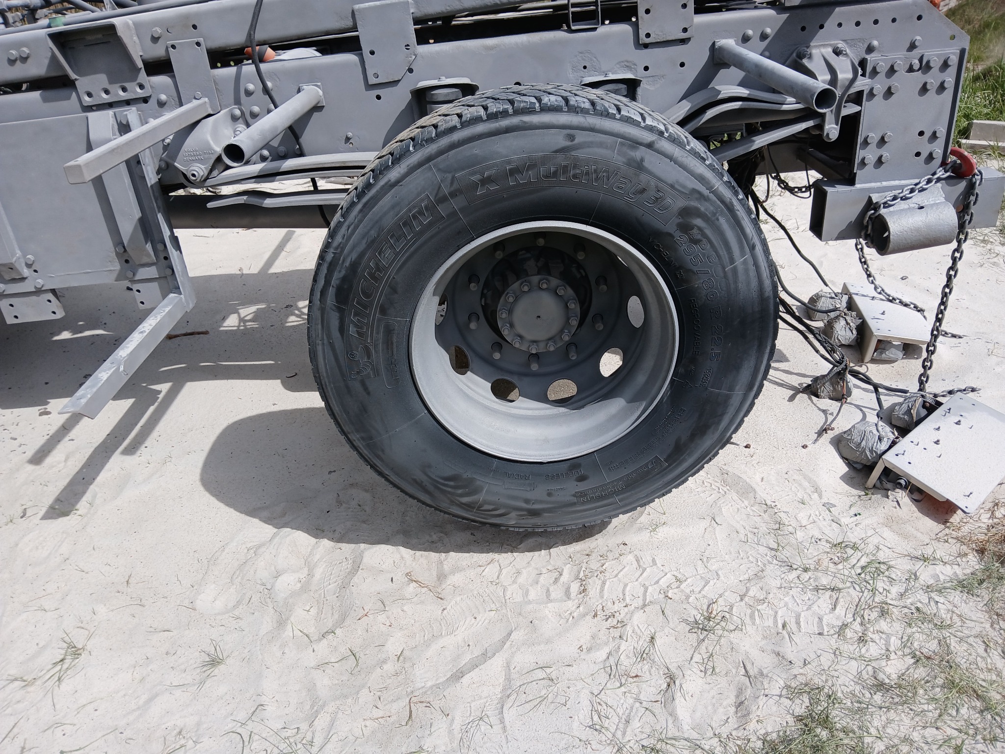 Szare podwozie ciężarówki z nową oponą Michelin X MultiWay 3D na piaszczystym podłożu. Widoczne ślady stóp i trawa. Elementy podwozia po piaskowaniu.