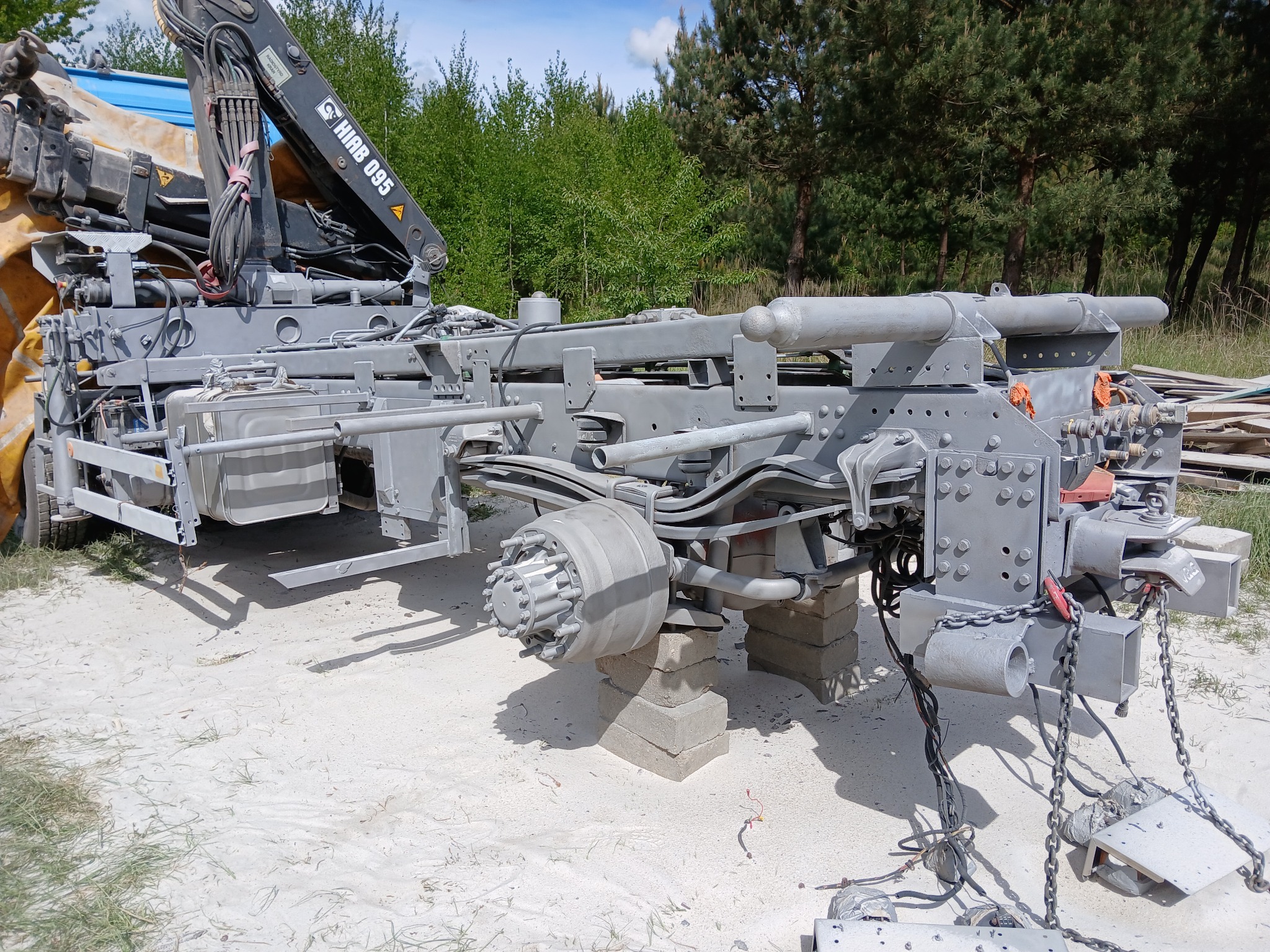 Szary podwozie ciężarówki z dźwigiem, podparte na betonowych blokach na piaszczystym podłożu. Widoczne elementy konstrukcyjne i mechaniczne. W tle zieleń drzew.