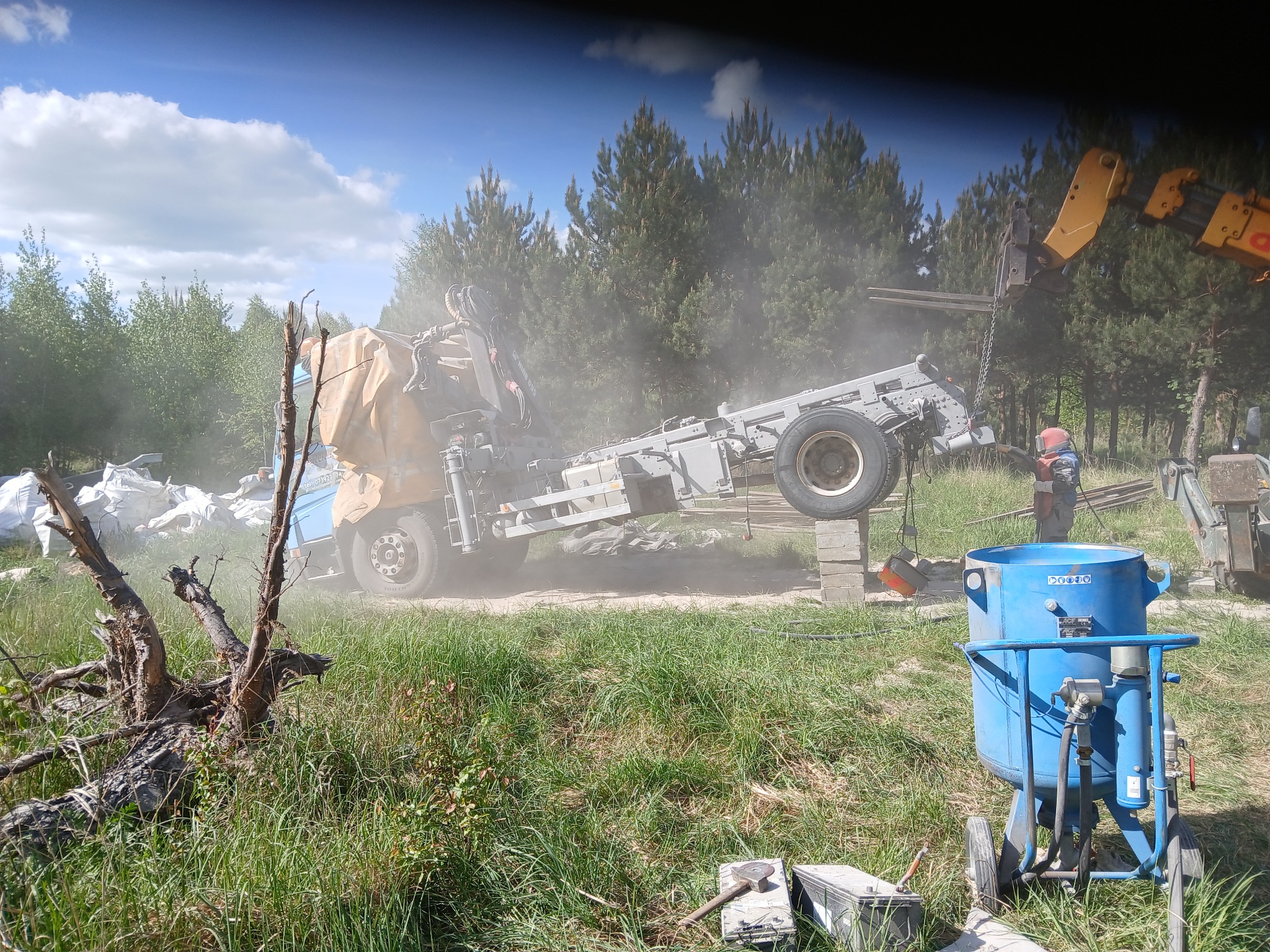 Piaskarka mobilna, błękitny zbiornik na kółkach, w trakcie pracy na zewnątrz, w tle drzewa i unoszący się pył. Widoczny fragment dźwigu i operator w kasku.