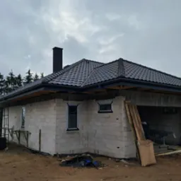 Nowy, grafitowy dach z kominem na surowym budynku z bloczków, z widocznymi rusztowaniami i materiałami budowlanymi. Pochmurne niebo w tle.