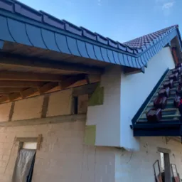 Widok na budynek w trakcie krycia dachu z użyciem dachówki ceramicznej. Część dachu pokryta dachówką karpiówką. Widoczne elementy konstrukcji dachu.