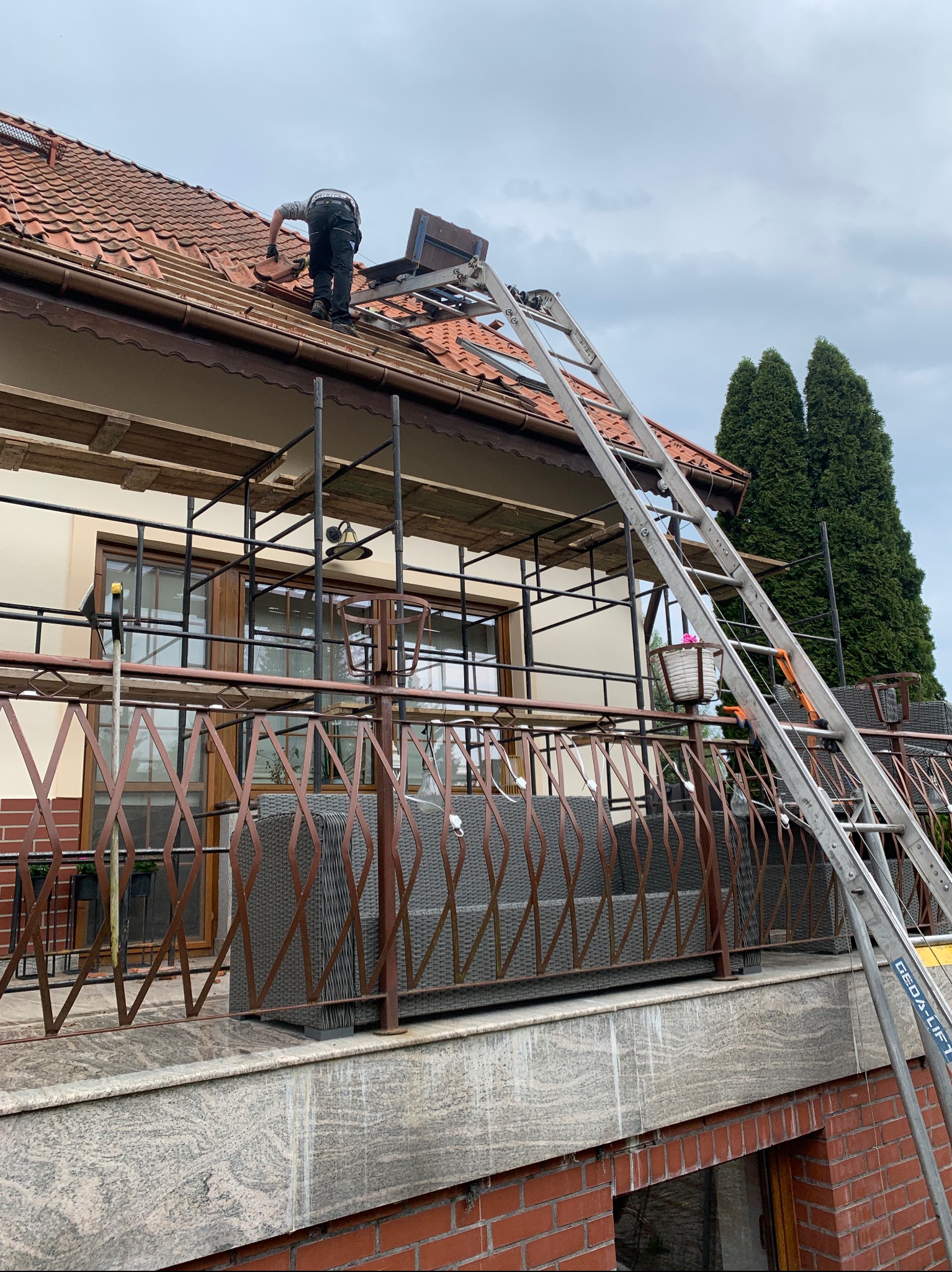 Dekarz wymienia dachówkę na dachu domu z użyciem windy dekarskiej. Widoczne rusztowanie i balkon z meblami ogrodowymi. Pochmurne niebo w tle.