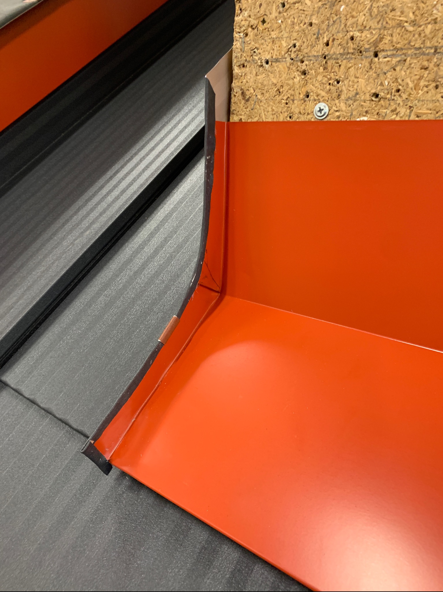 Pomarańczowa obróbka blacharska przy ciemnoszarym dachu, widoczna płyta OSB z wkrętem. Detal wykonania orynnowania dachu w zbliżeniu.